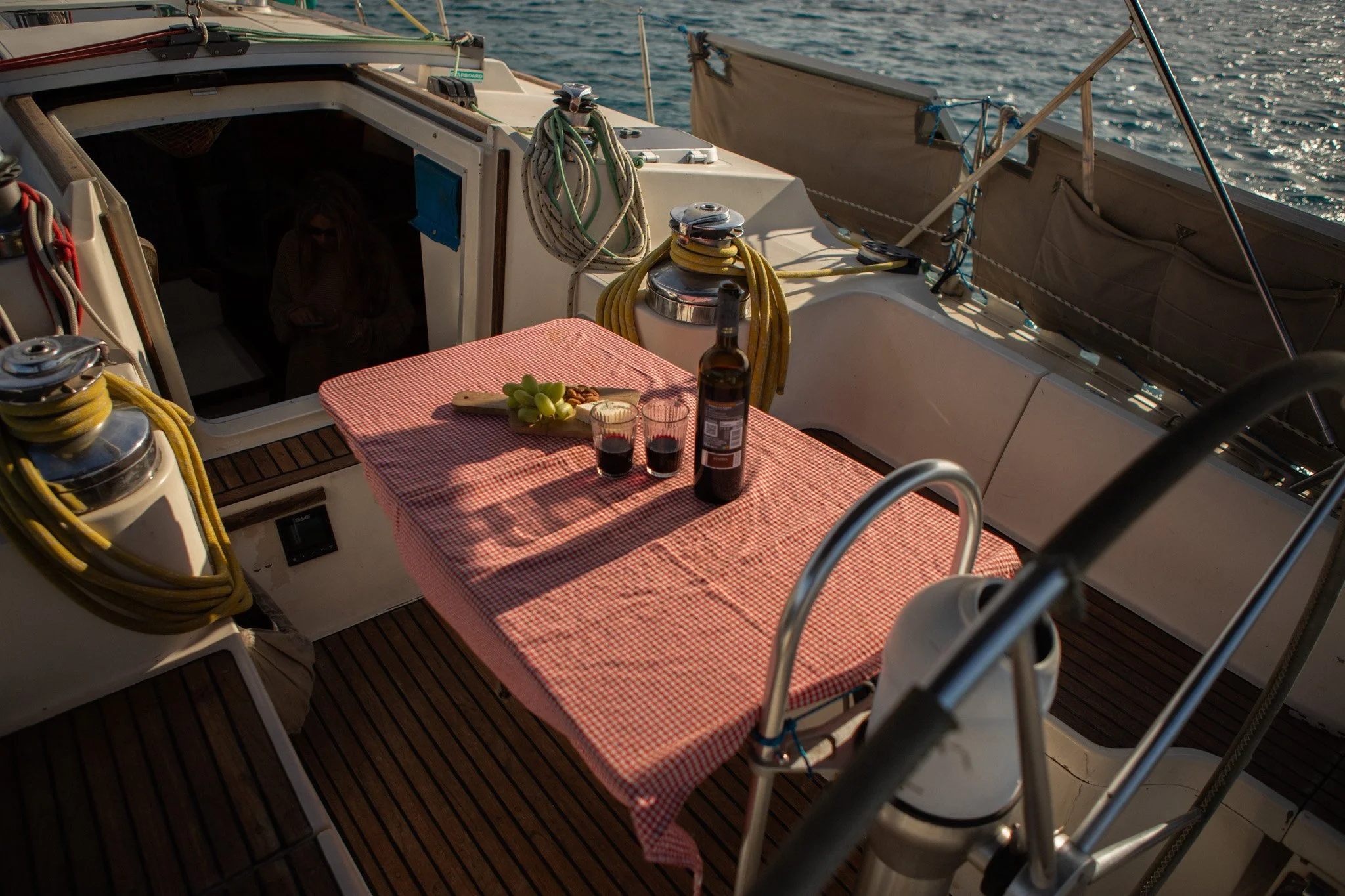 Mesa con mantel de cuadros rojos y blancos, con una botella de vino y dos copas con vino tinto, en la cubierta de un yate, rodeada de cuerdas, winches y barandillas, con el mar de fondo.