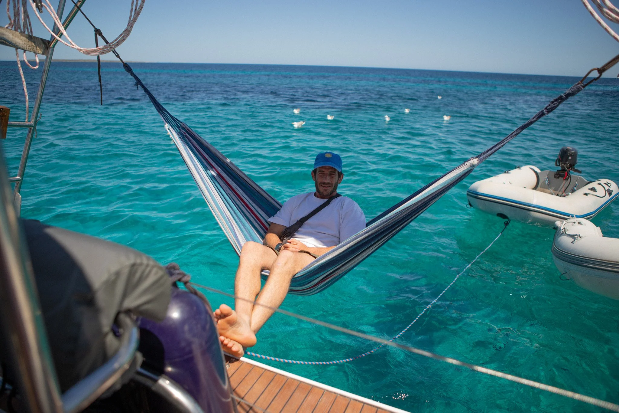 Persona descansando en una hamaca en la cubierta de un barco, con el mar azul de fondo y una lancha inflable a su lado.
