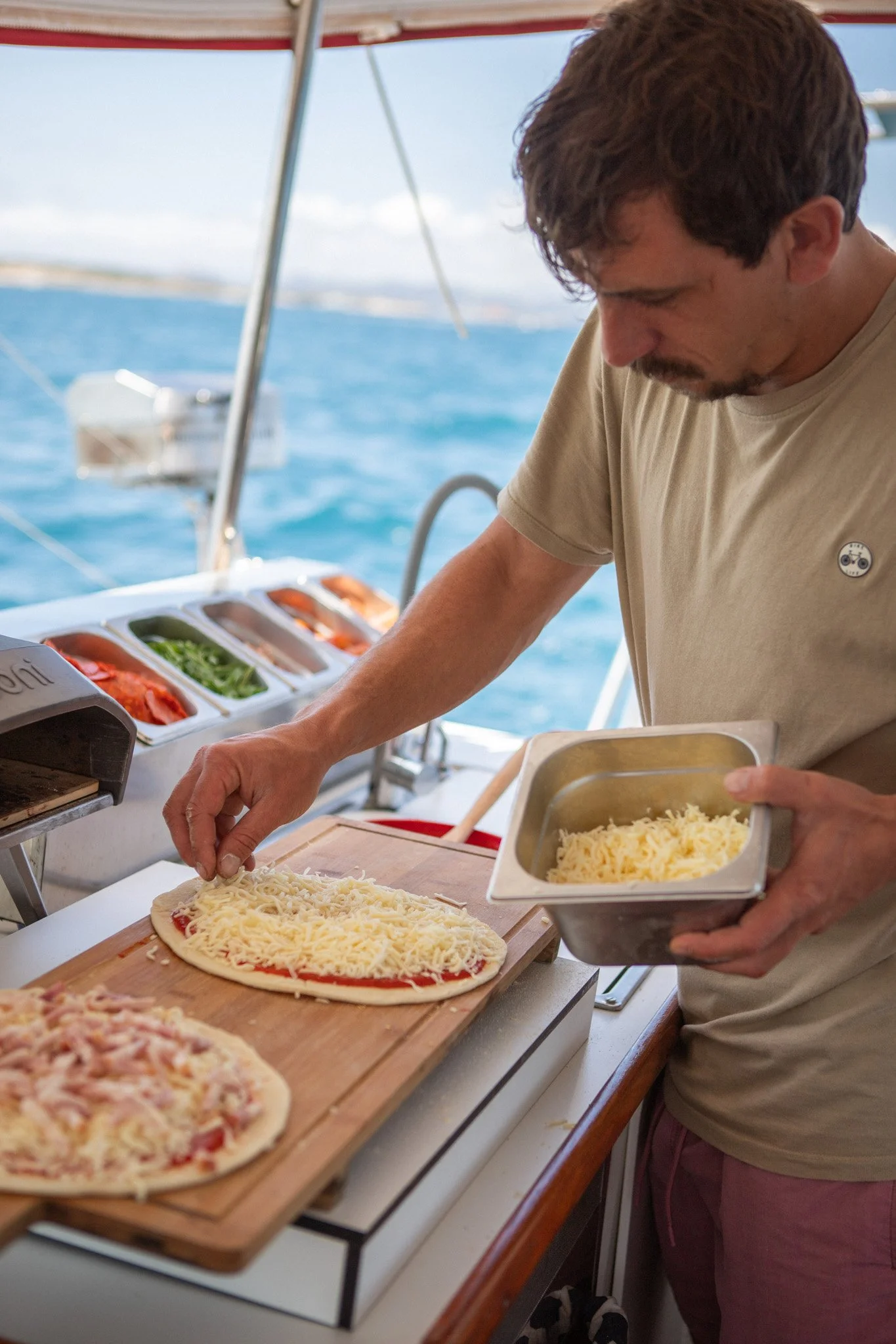Persona preparando una pizza con queso rallado en una cocina al aire libre en un barco