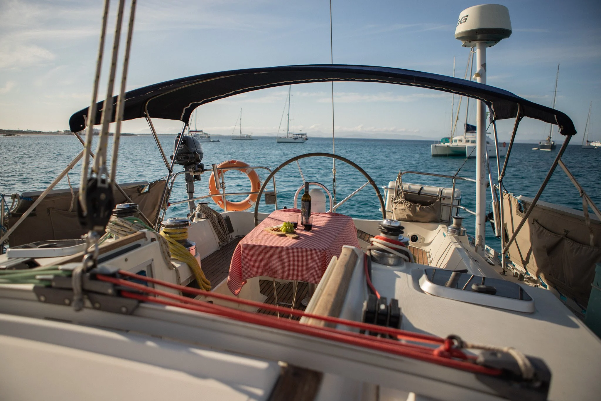 Vista desde la cabina de un velero con mesa y sillas, mostrando el mar con varios barcos anclados en un día soleado