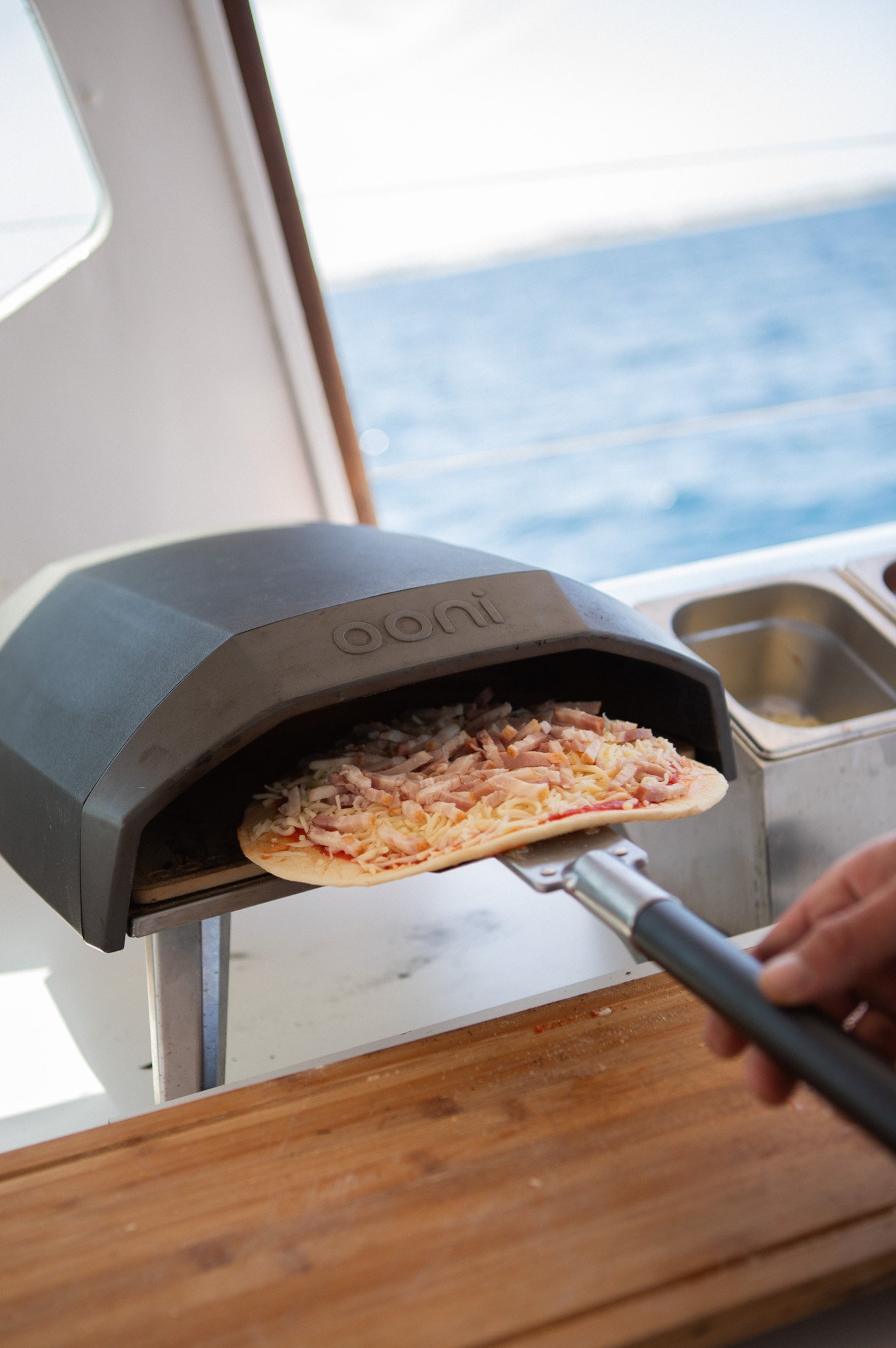 Persona colocando una pizza en un horno de pizza casero con un entorno que parece ser en el mar, con vista del océano y cielo azul, en una decoración moderna con mesa de madera.