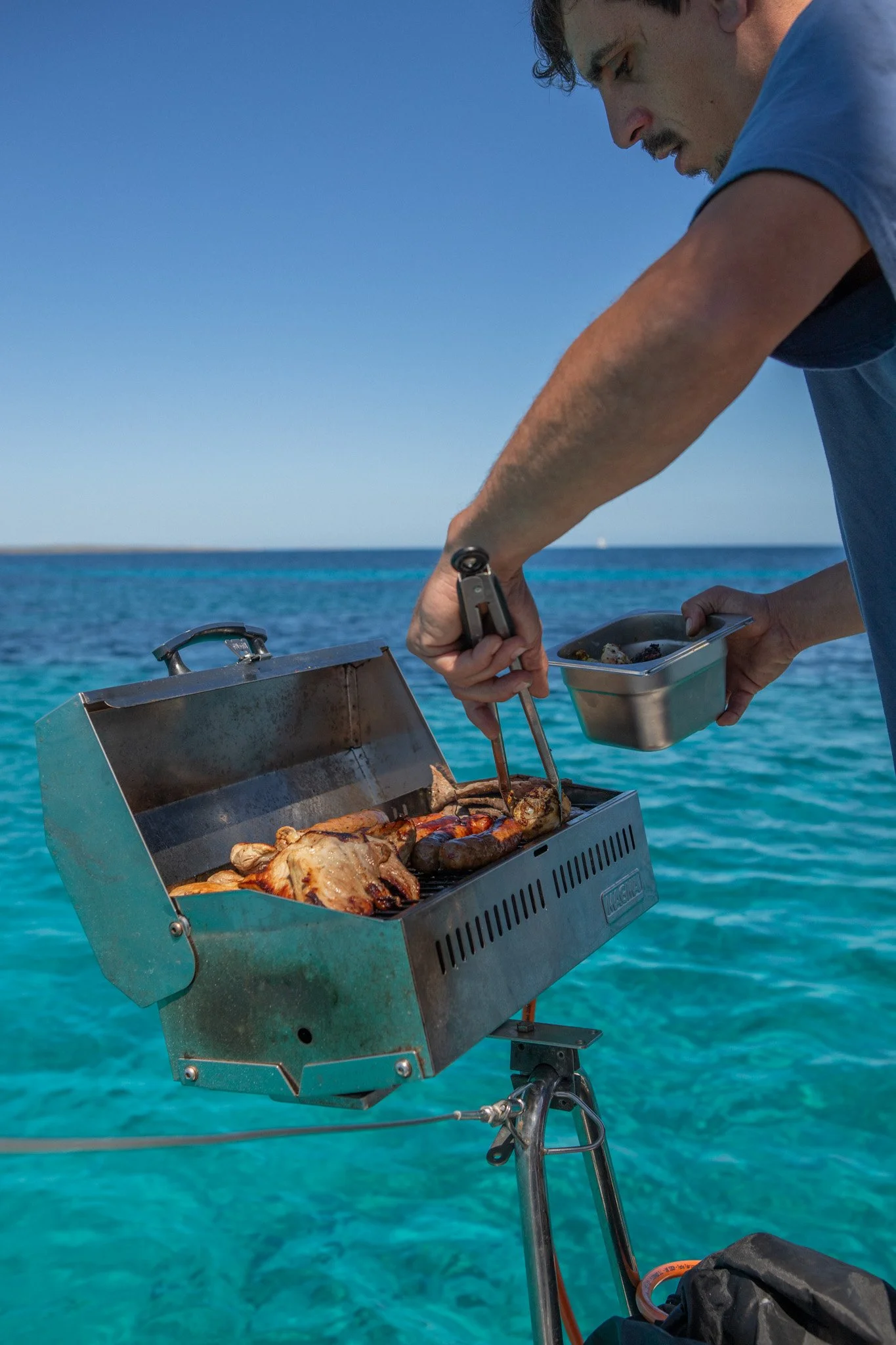 Persona cocinando carne en una parrilla portátil en un ambiente junto al mar, con agua azul clara y cielo despejado.