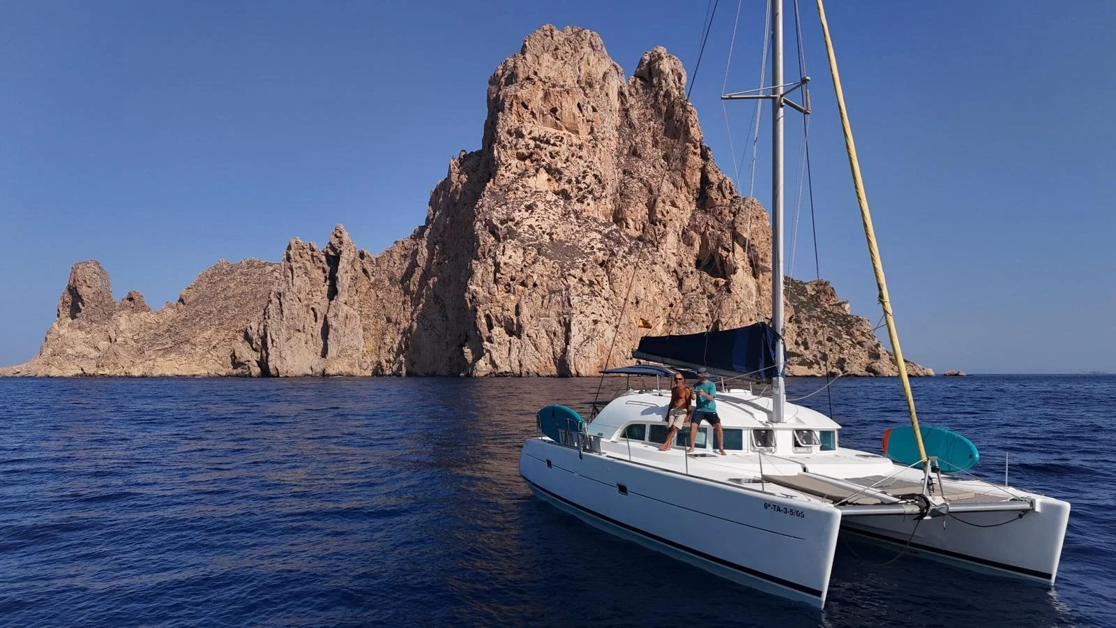 Barco catamarán en el mar, con dos personas en la cubierta, frente a una formación rocky en un día soleado y despejado.