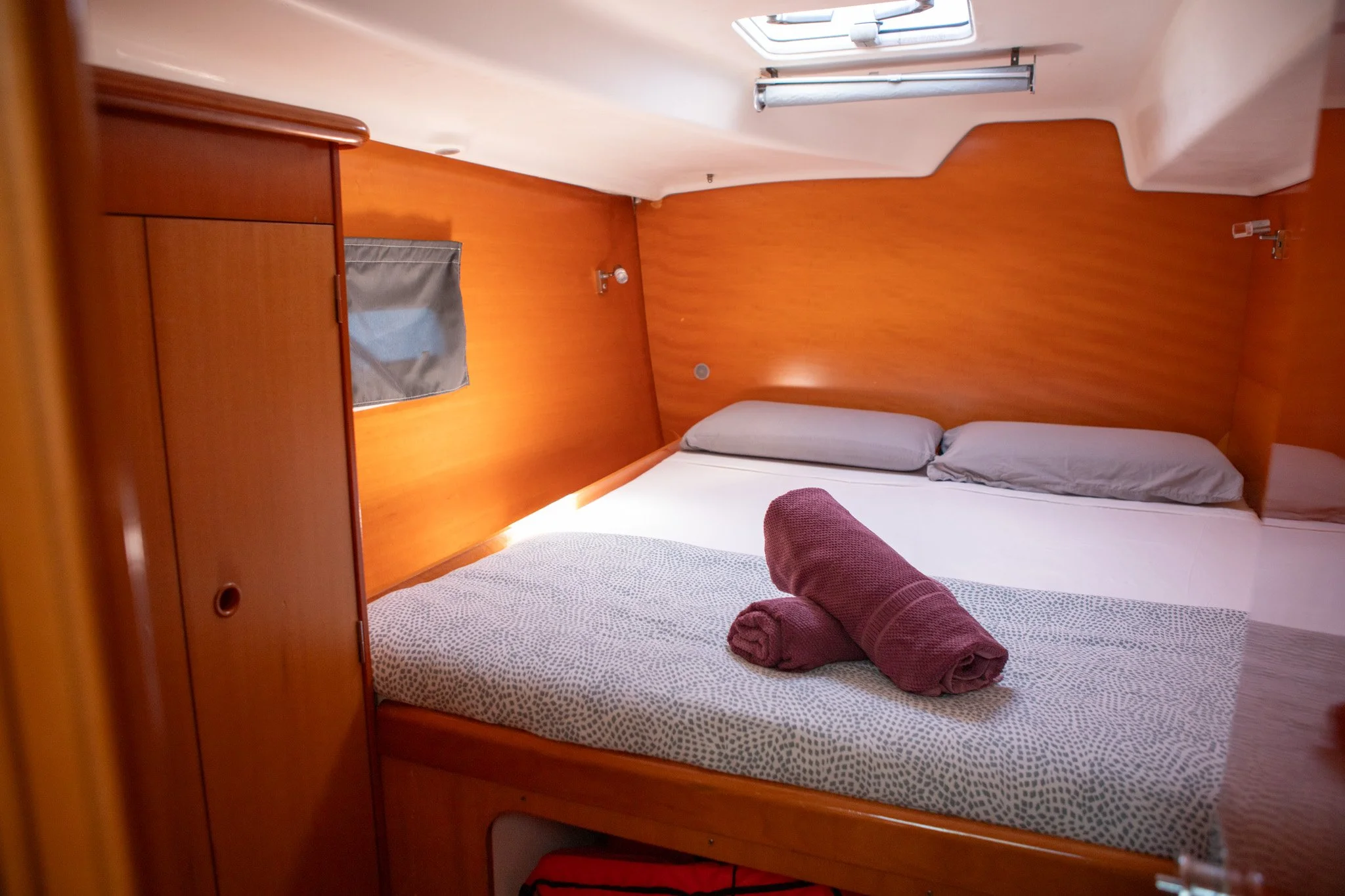 Cama en una cabina de barco con almohadas gris y sábanas blancas, cortinas de tela gris, y una ventana en el techo que deja entrar luz.