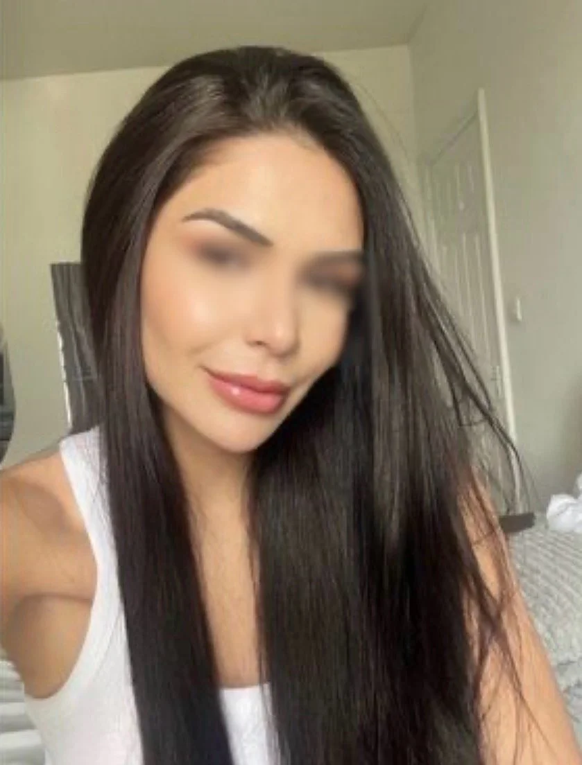 A woman with long, straight dark hair smiling at the camera, wearing a white sleeveless top, in a room with beige walls and a door in the background.