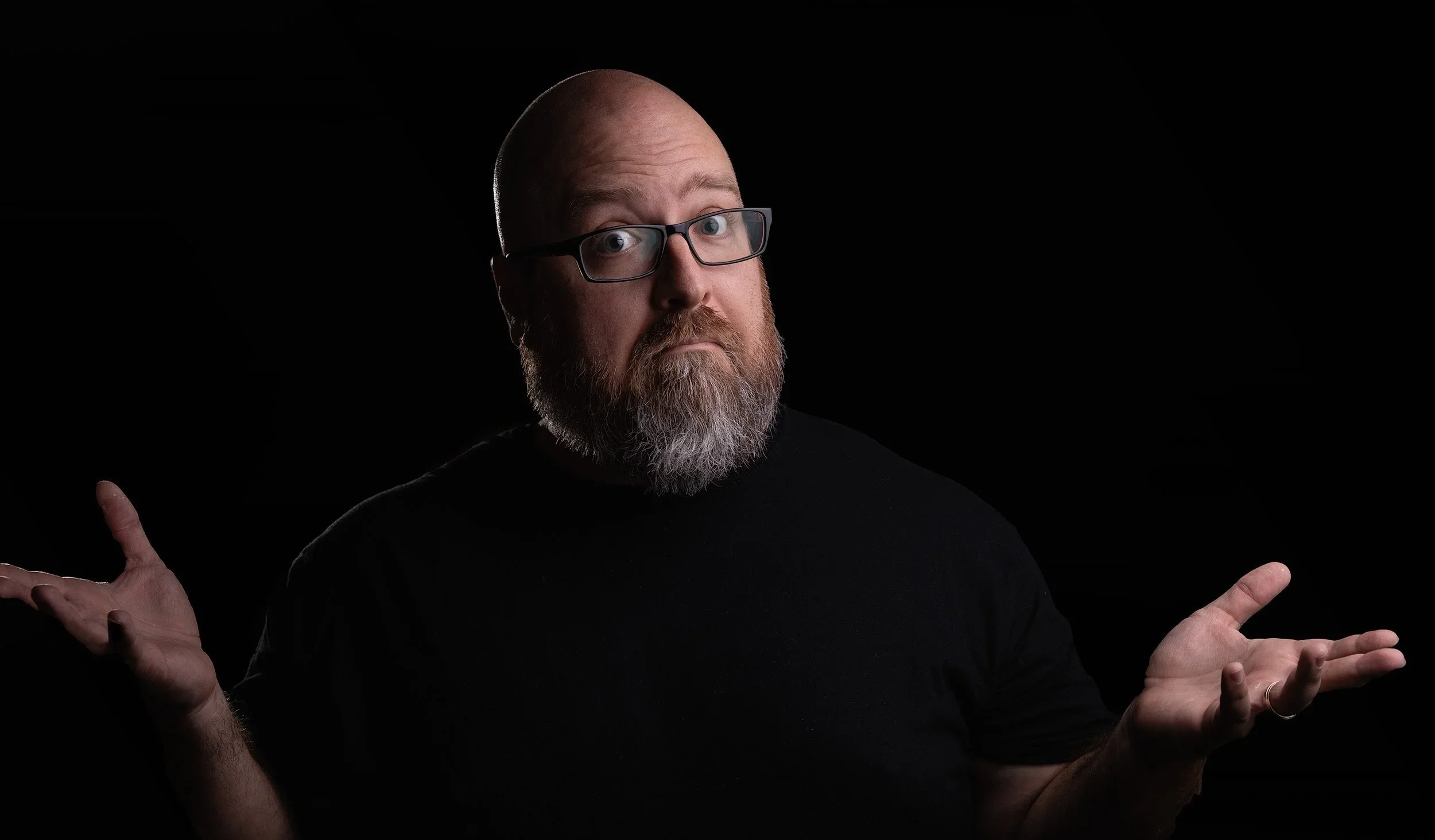 A man with glasses, a beard, and a bald head, wearing a black shirt, looking at the camera with raised shoulders and palms facing upward against a black background.