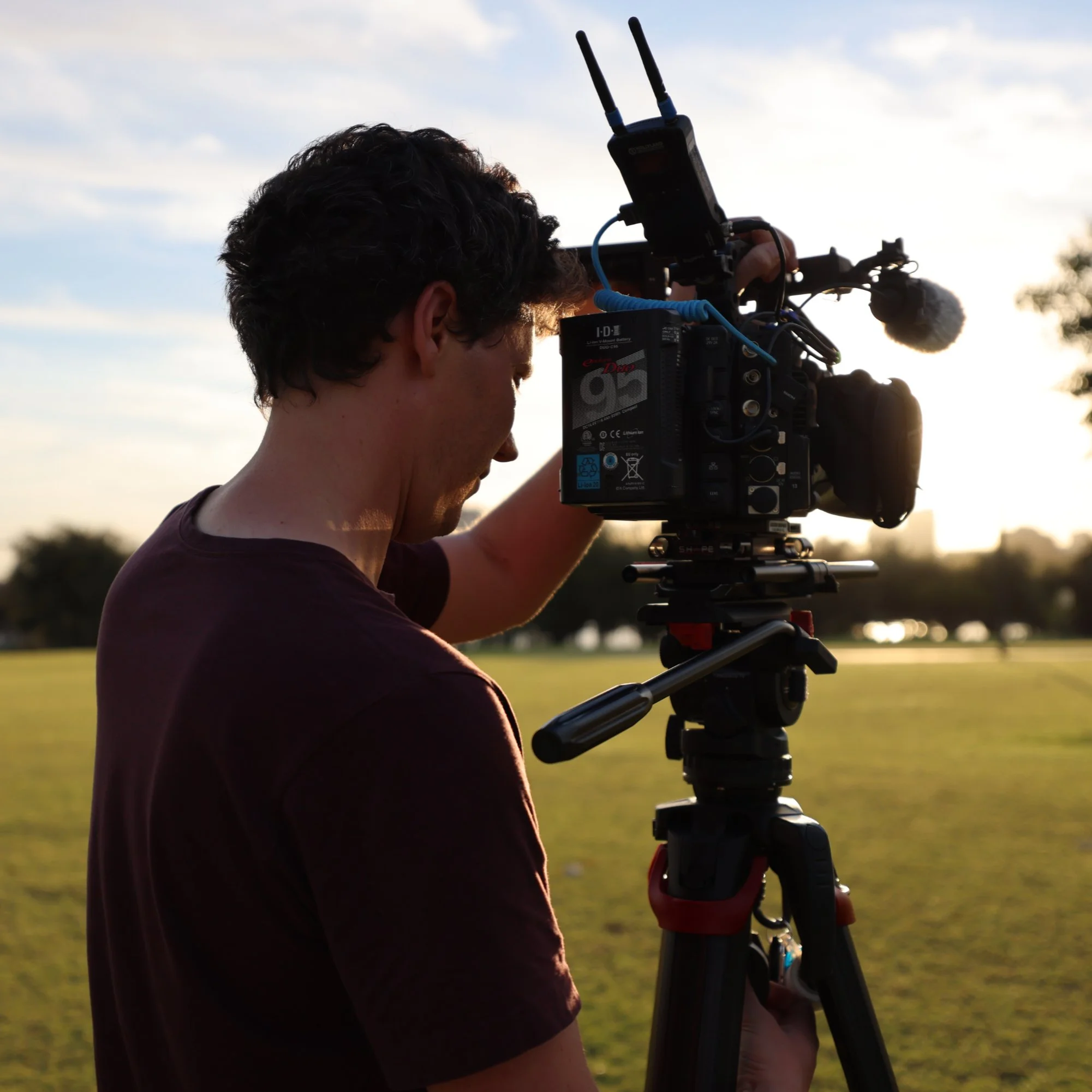 A person with dark curly hair operating a professional video camera on a tripod outdoors during sunset.