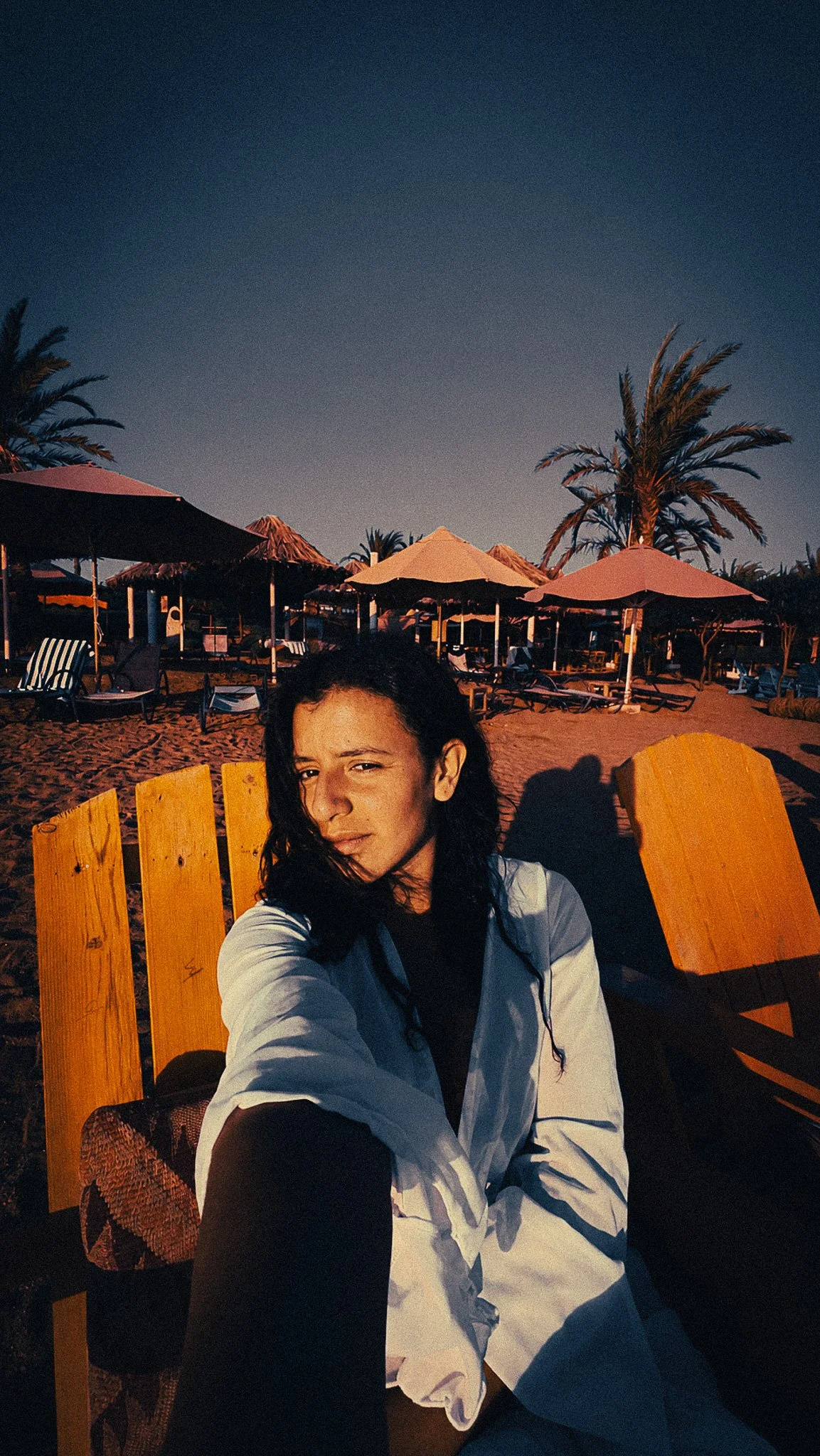 Woman sitting on an orange beach chair in Aqaba, with palm trees, beach umbrellas and warm Red Sea light in the background.
