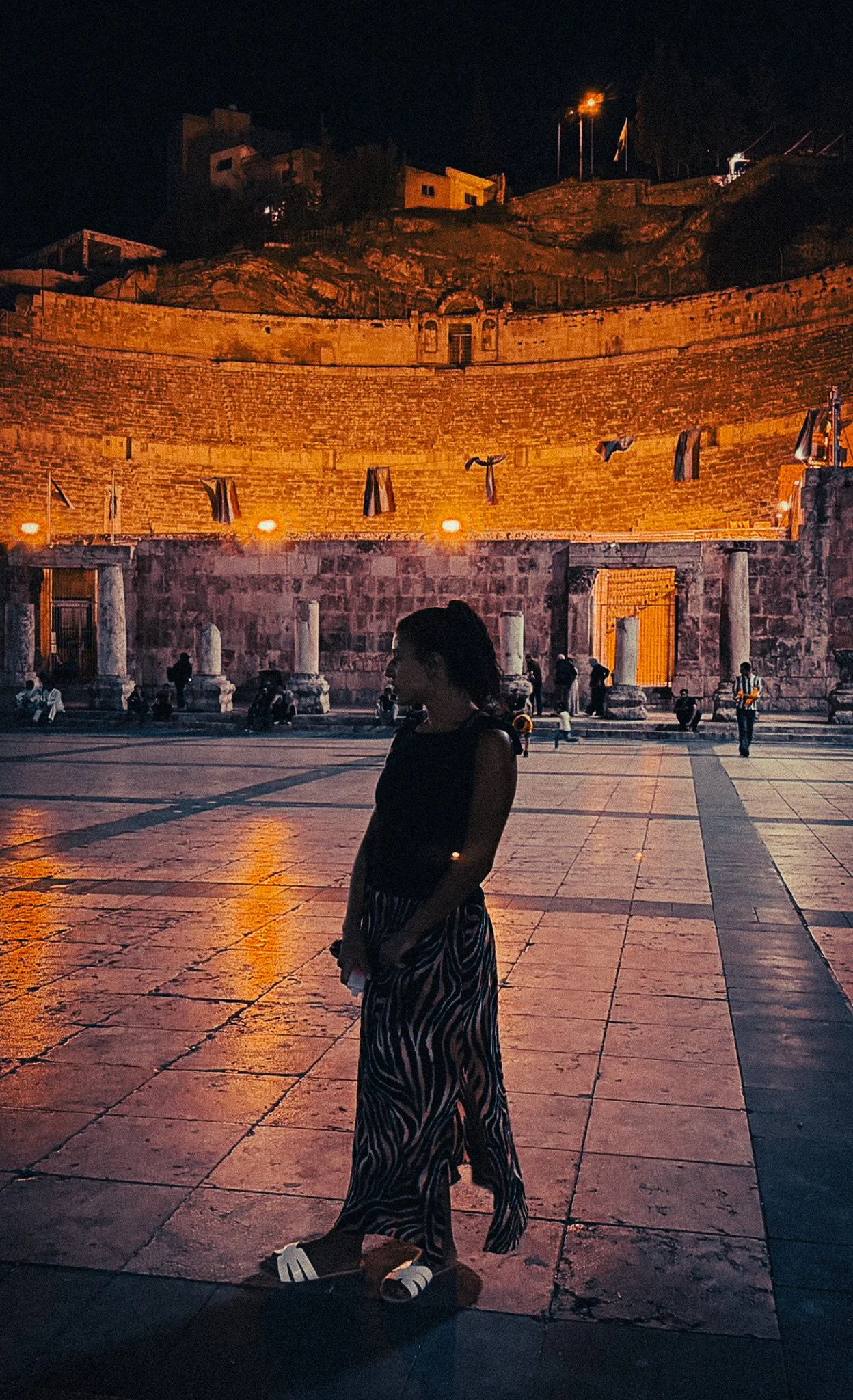 A woman standing in front of the Roman Theatre in Amman at night, with warm lights illuminating the ancient stone seats.