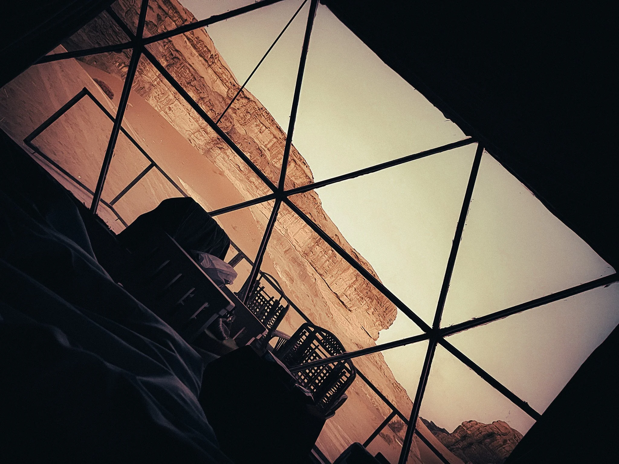 View from inside a Wadi Rum bubble tent, showing its geometric glass frame, desert cliffs and outdoor chairs.