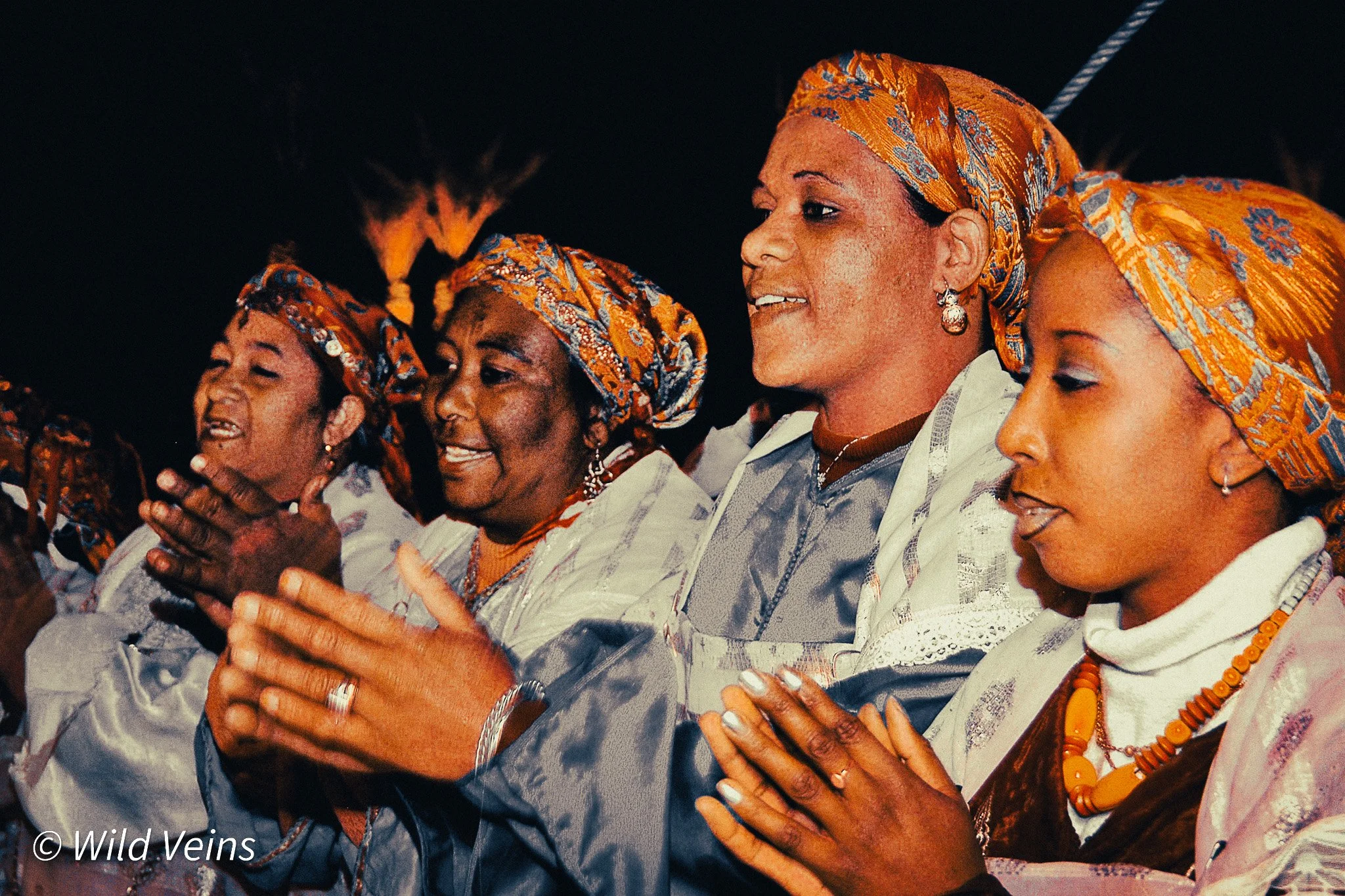 A group of women wearing vibrant Moroccan traditional dress, orange patterned headscarves, jewellery, and blue and pink fabrics clap together during an evening music performance.