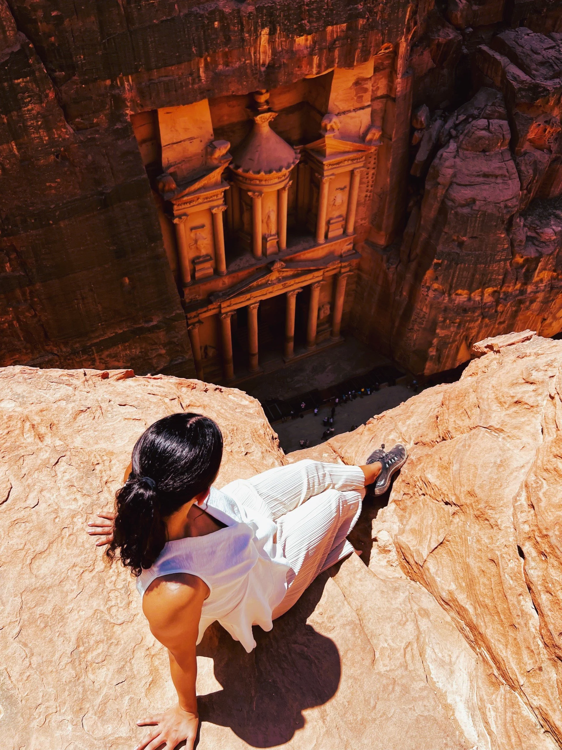 A woman in a white dress sitting on a rock ledge, overlooking the ancient city of Petra, Jordan, with rock-cut architecture visible in the canyon below, during daytime.