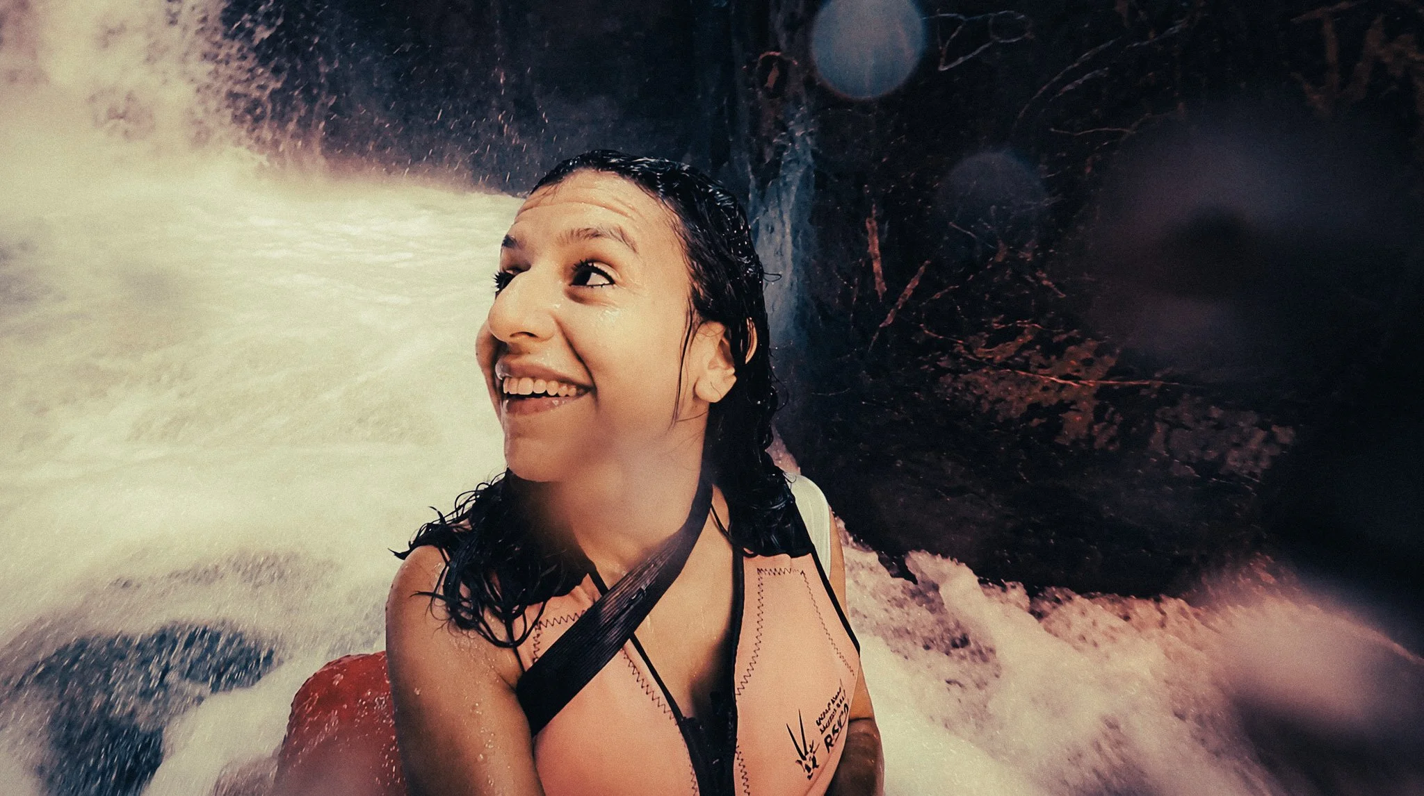 Woman looking up and smiling beside rushing white water and dark canyon walls during the Wadi Mujib Siq Trail.