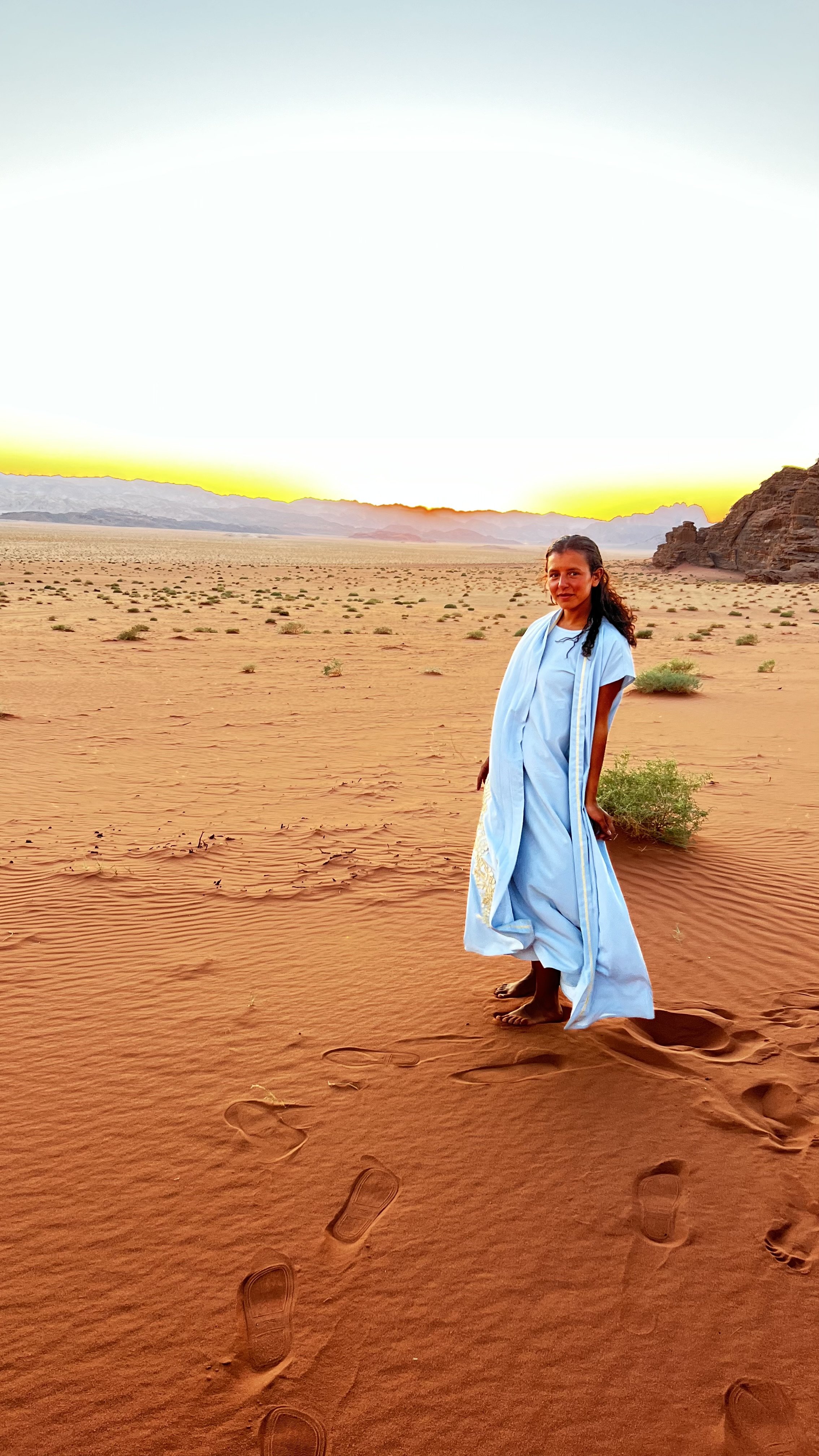 A woman in a blue dress standing barefoot on orange desert sand at sunset, with sand dunes and rocky formations in the background.