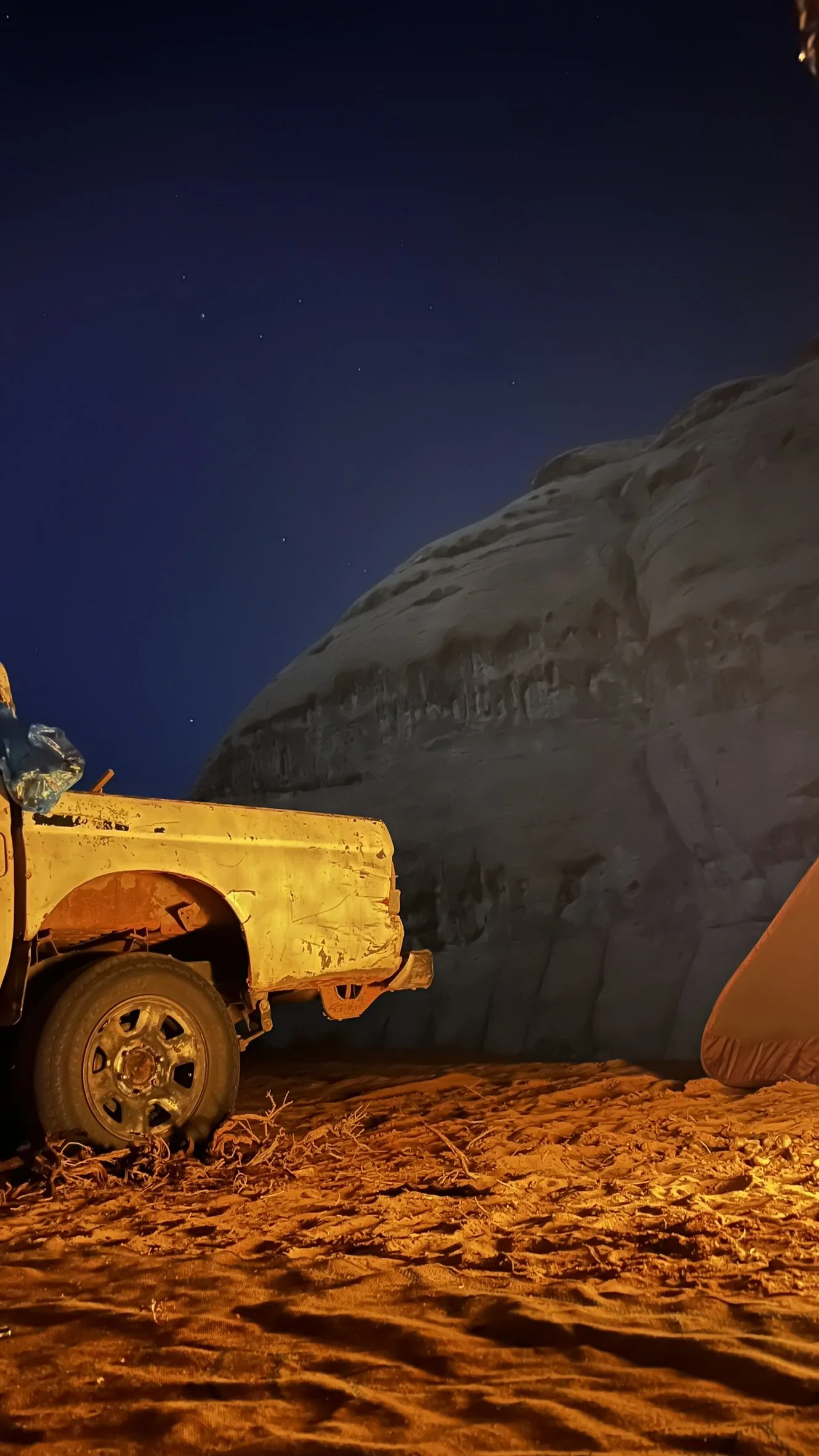 A yellow jeep parked beside tents under the stars in Wadi Rum desert at night, with red sand and rock formations around the camp.