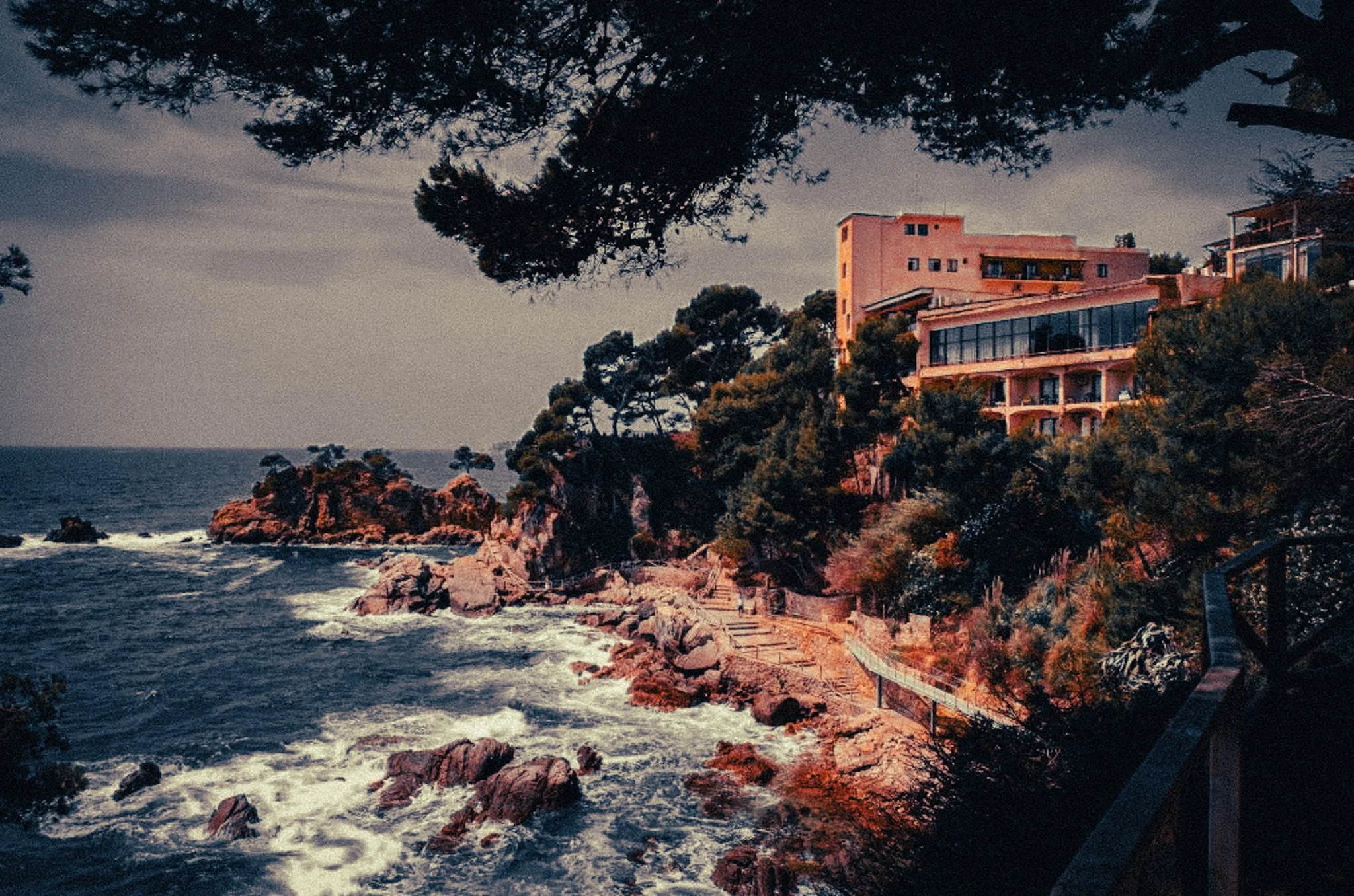A seaside house on a rocky cliff surrounded by trees, with waves crashing below on a cloudy day.