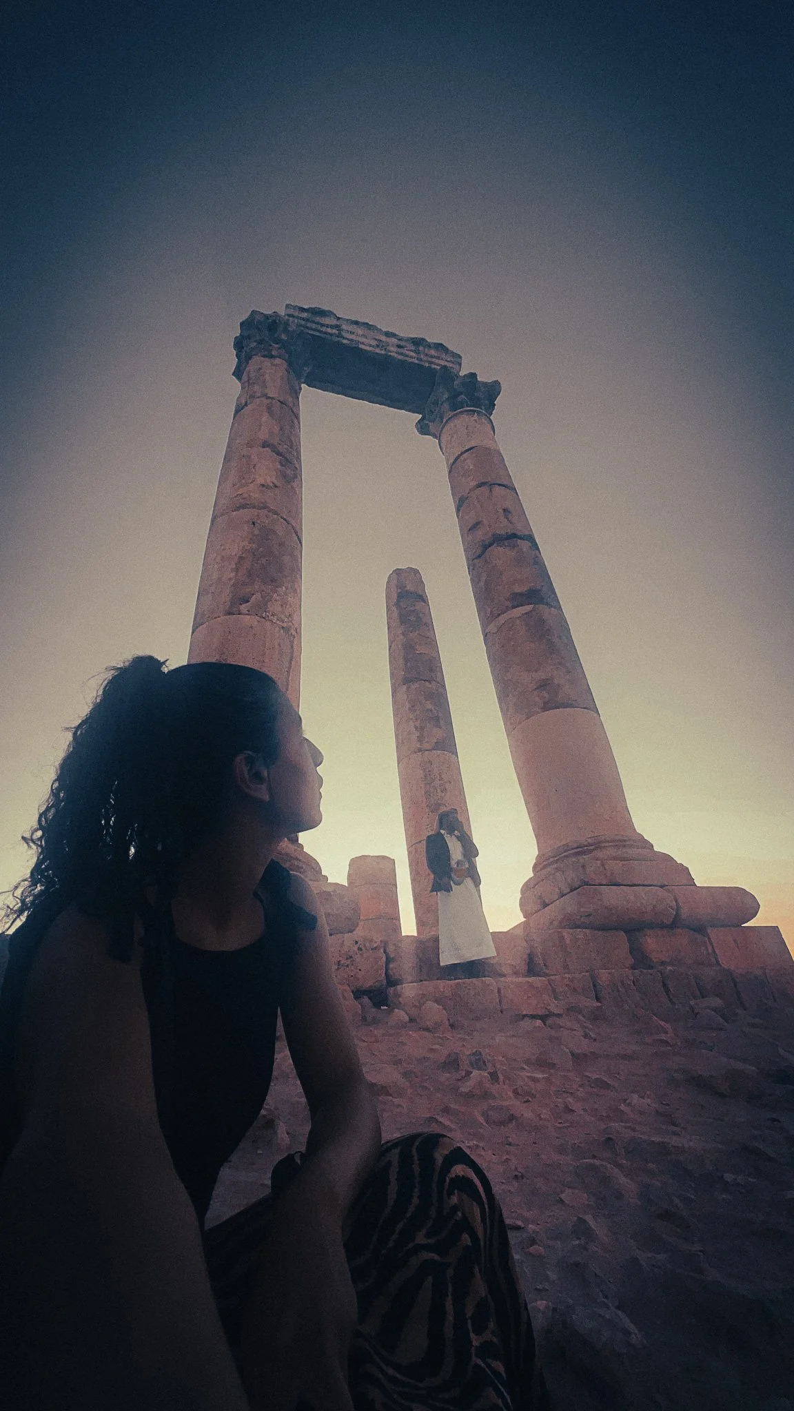 Woman sitting in silhouette at the Amman Citadel, looking toward tall ancient Roman columns at sunset.