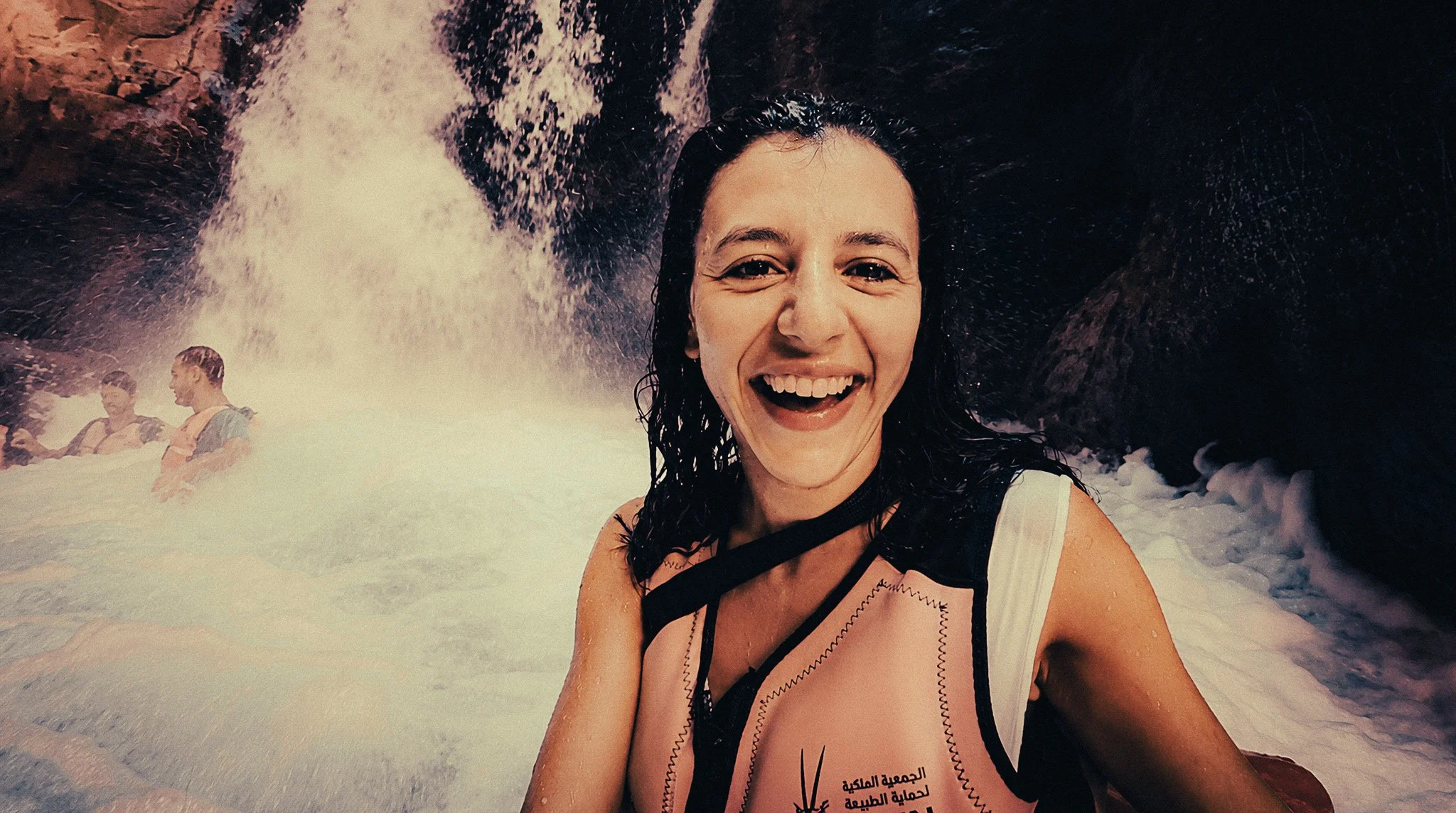 Woman smiling in a life jacket beneath a powerful waterfall while canyoning through Wadi Mujib in Jordan.