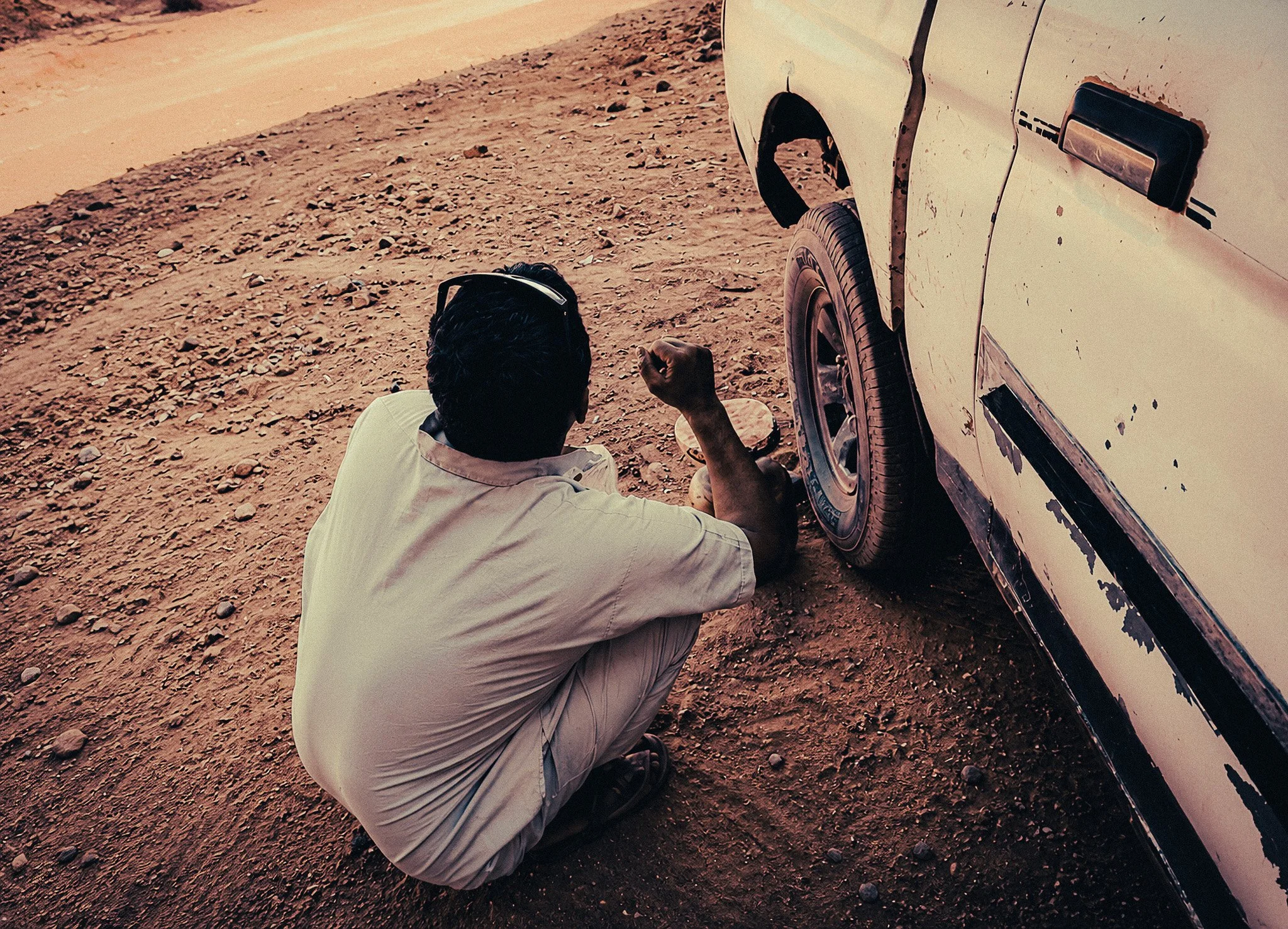 Bedouin man crouching beside a weathered white jeep during a stop in the rocky Wadi Rum desert.