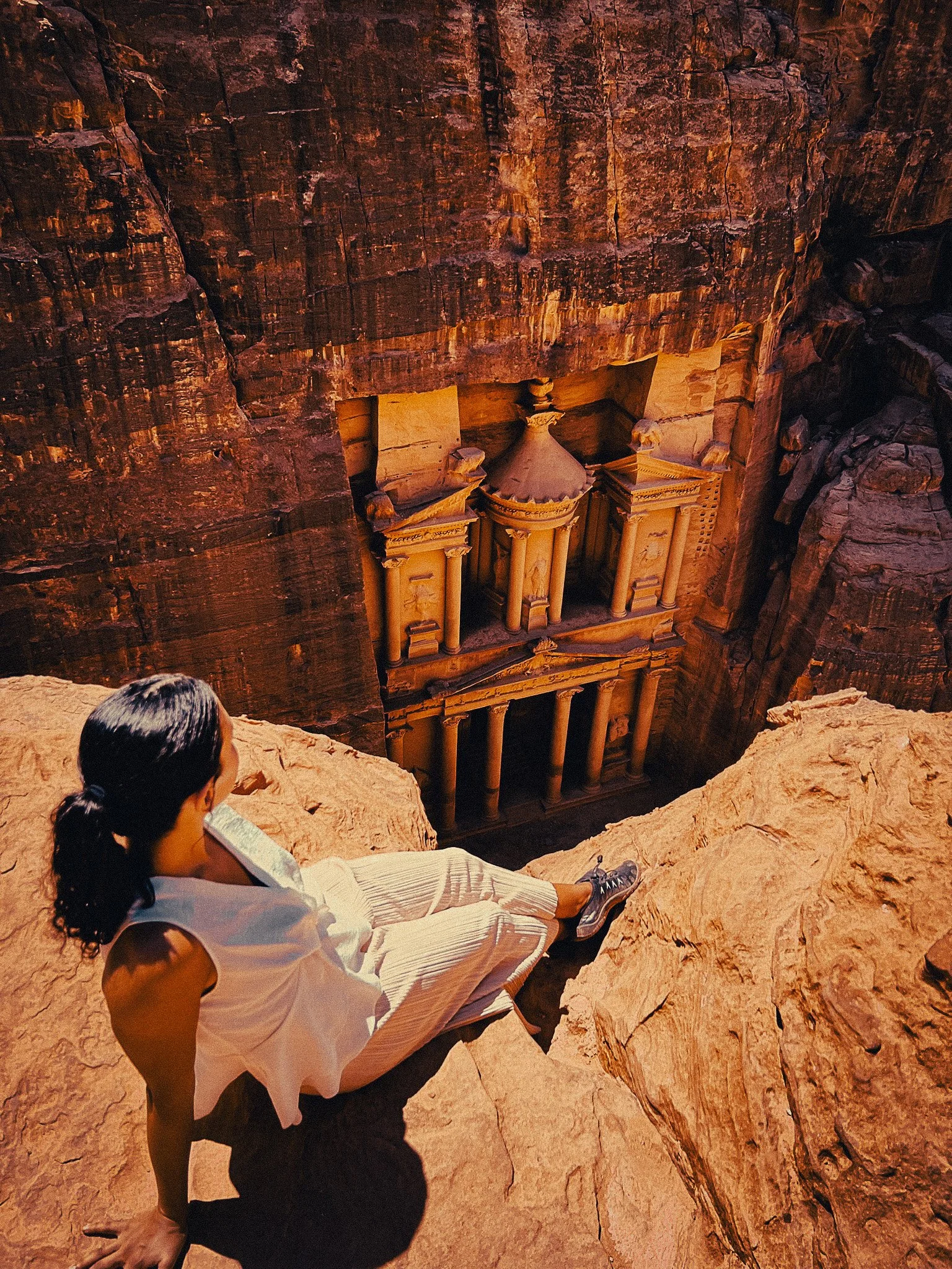 Woman sitting on a high viewpoint above Petra’s Treasury, looking down at the ancient carved façade between sandstone cliffs.