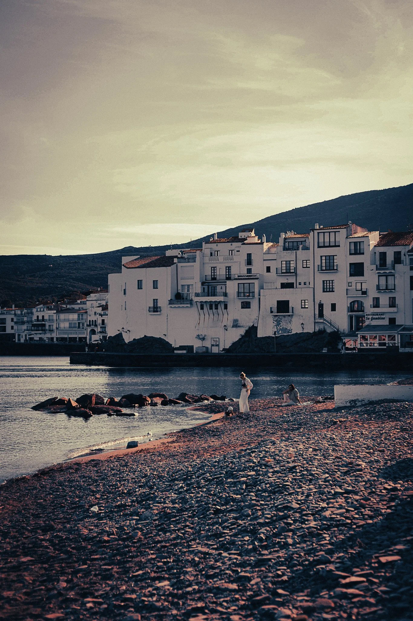 Cadaqués waterfront with white buildings beside a rocky beach, calm water and dark hills in the background on the northern Costa Brava.