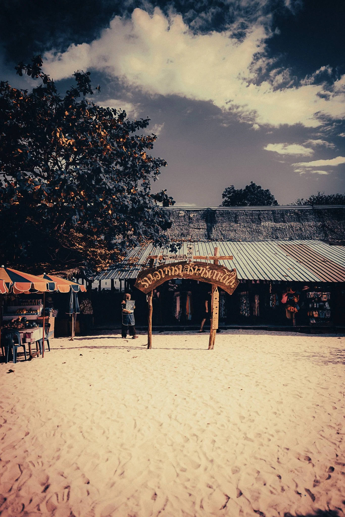 Beach entrance on Koh Bon near Rawai, Phuket, with sand, trees and a wooden Thai sign.