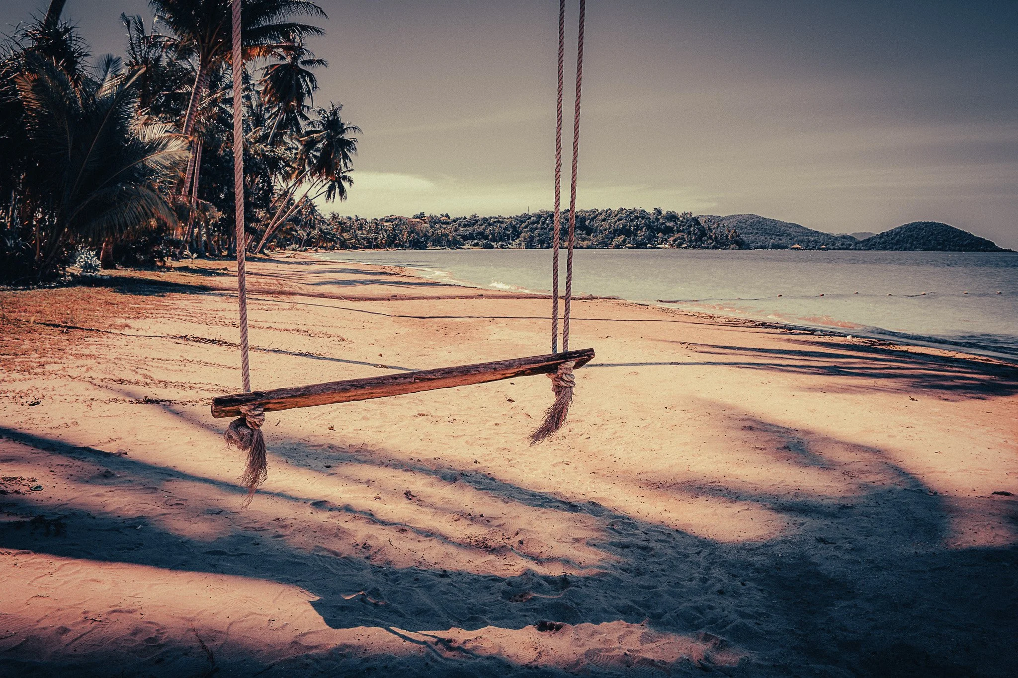 Empty wooden swing on a quiet beach in Koh Yao Yai, Thailand, with palm trees and calm sea.