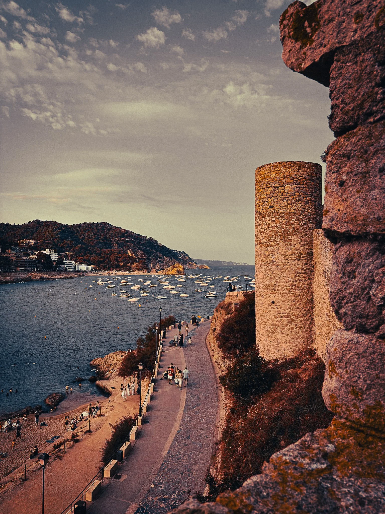 View from Tossa de Mar’s old stone walls over the bay, with a round medieval tower, rocky cliffs, boats in the water and the Costa Brava coastline in the distance.