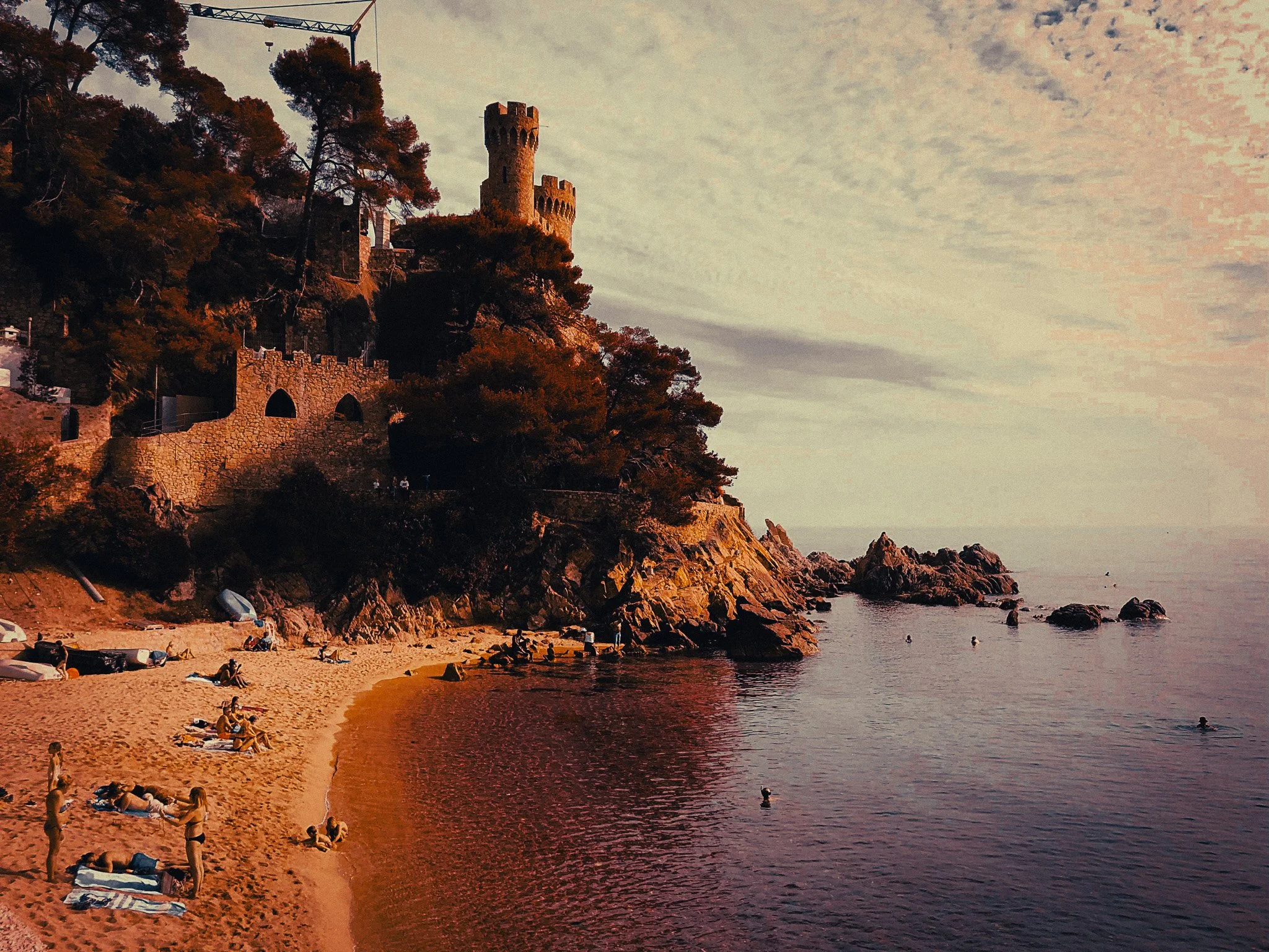 Small sandy cove on the Costa Brava with swimmers in calm water, rocky cliffs, pine trees and a stone building above the beach.