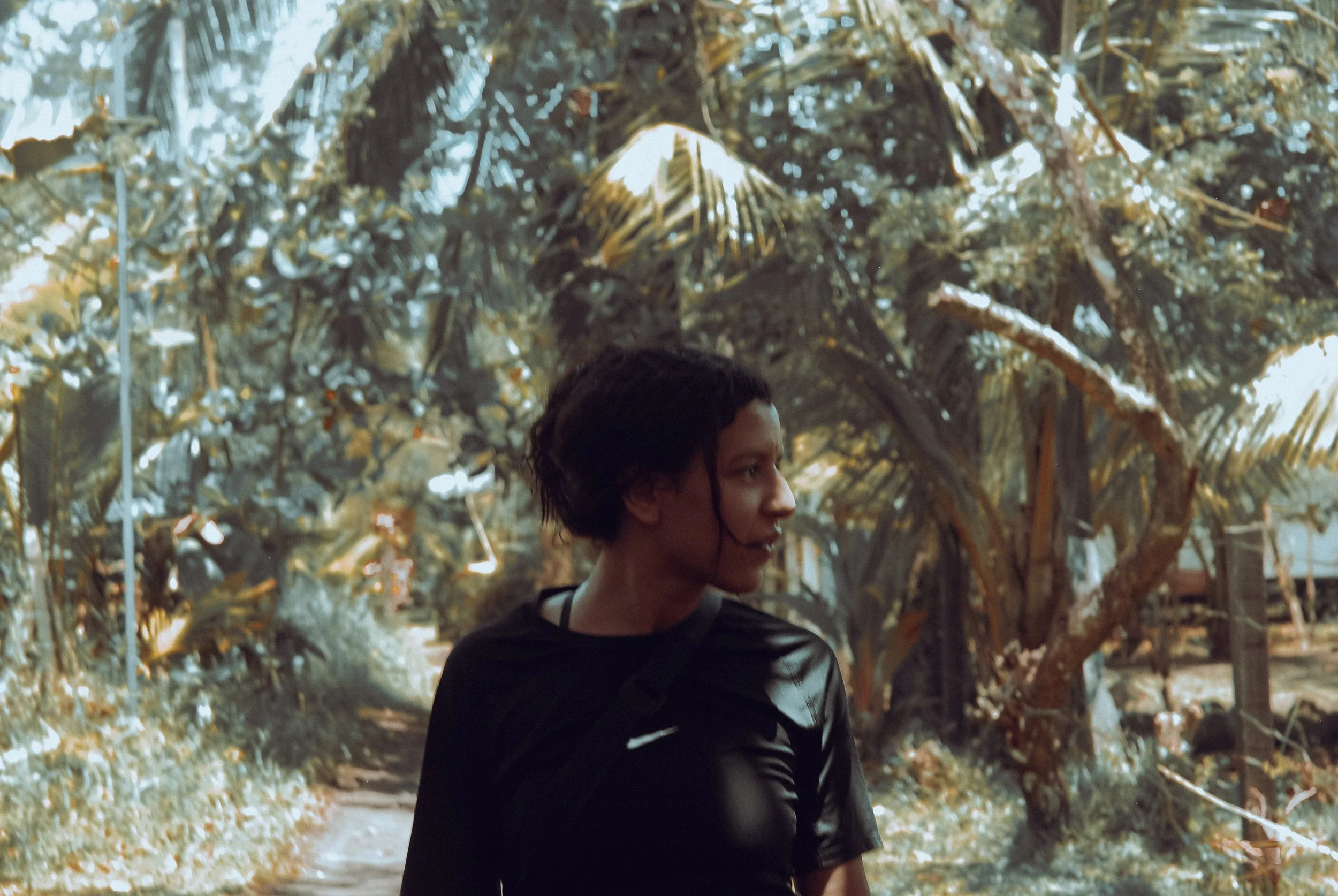 A person with short curly hair and a black shirt standing outdoors in a lush green garden or park area.