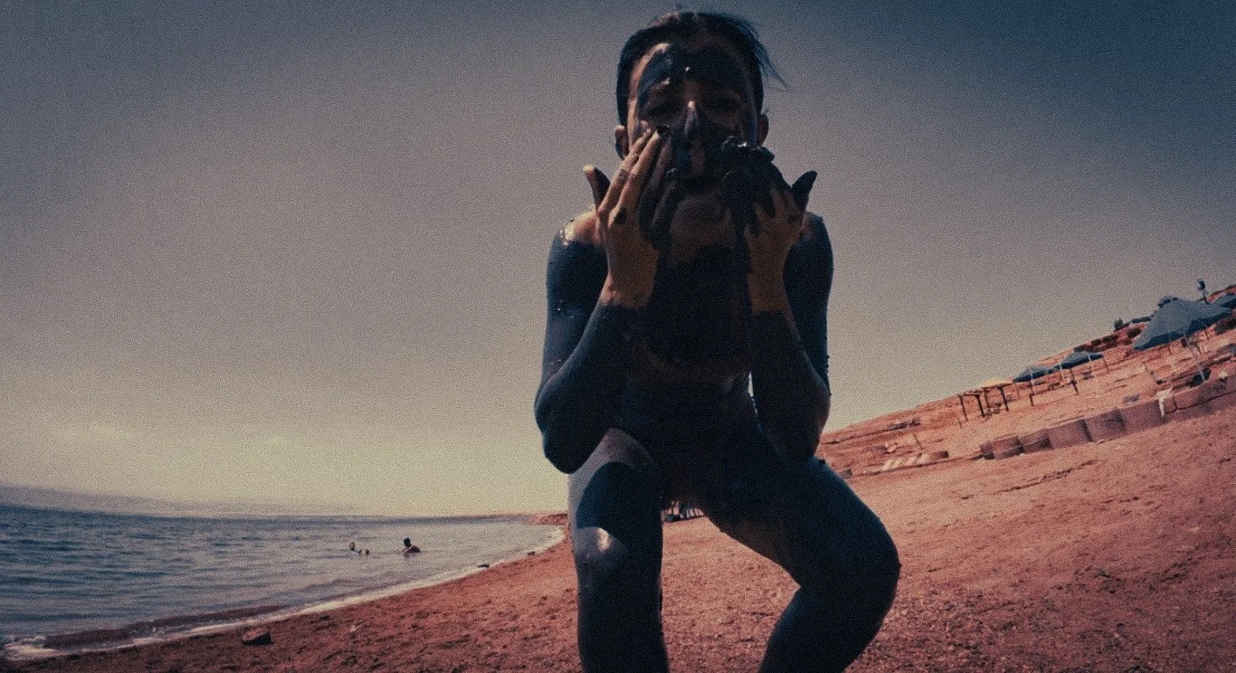 A traveller covered in Dead Sea mud sitting near the shore in Jordan, with the salty water behind them.