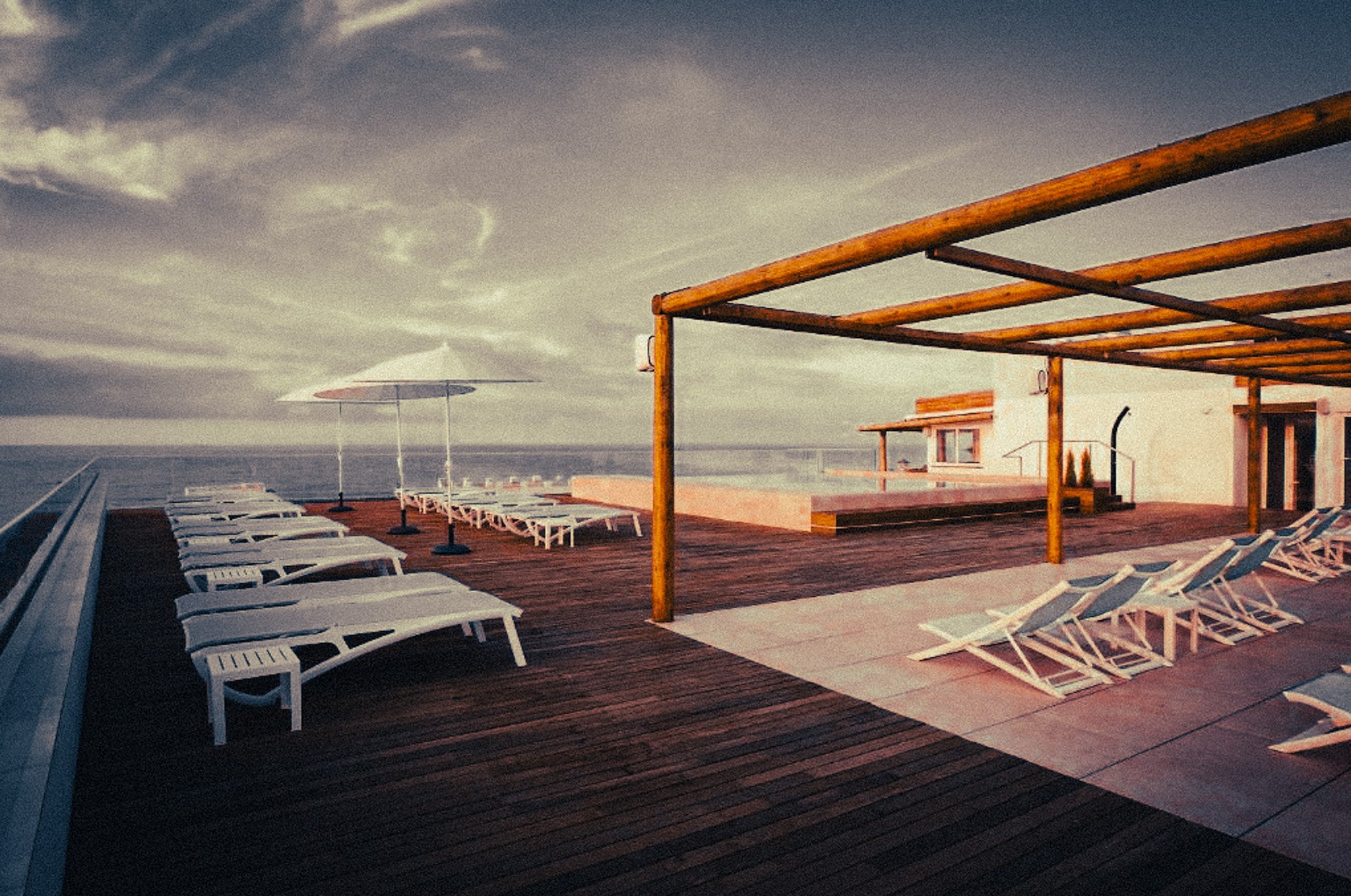 A rooftop deck with white lounge chairs, a wooden pergola structure, a large umbrella, and a section of an ocean view in the background at sunset or dusk.