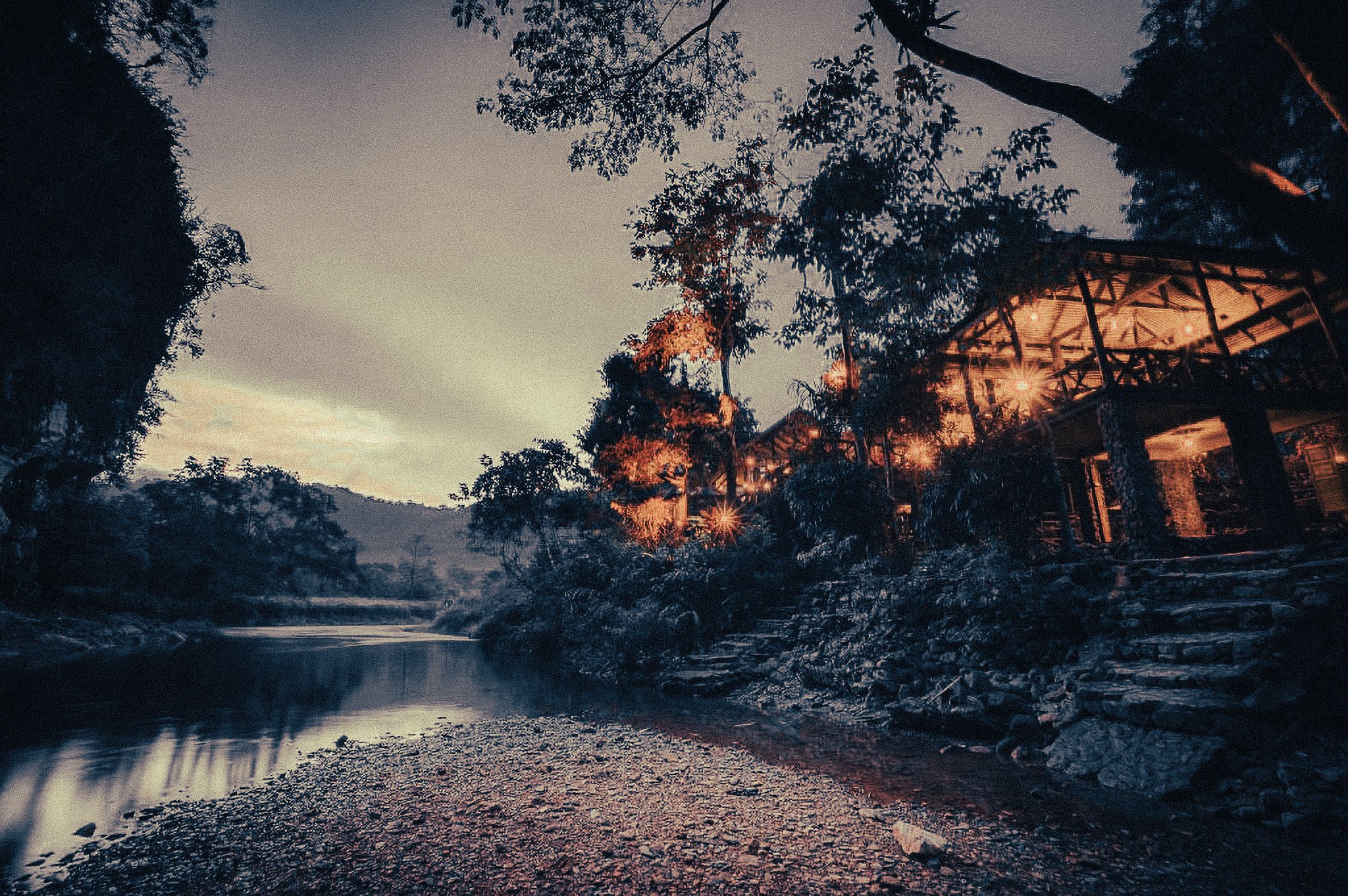 Riverside jungle lodge in Khao Sok, Thailand, glowing with warm lights at dusk beside a calm river and forested limestone hills.