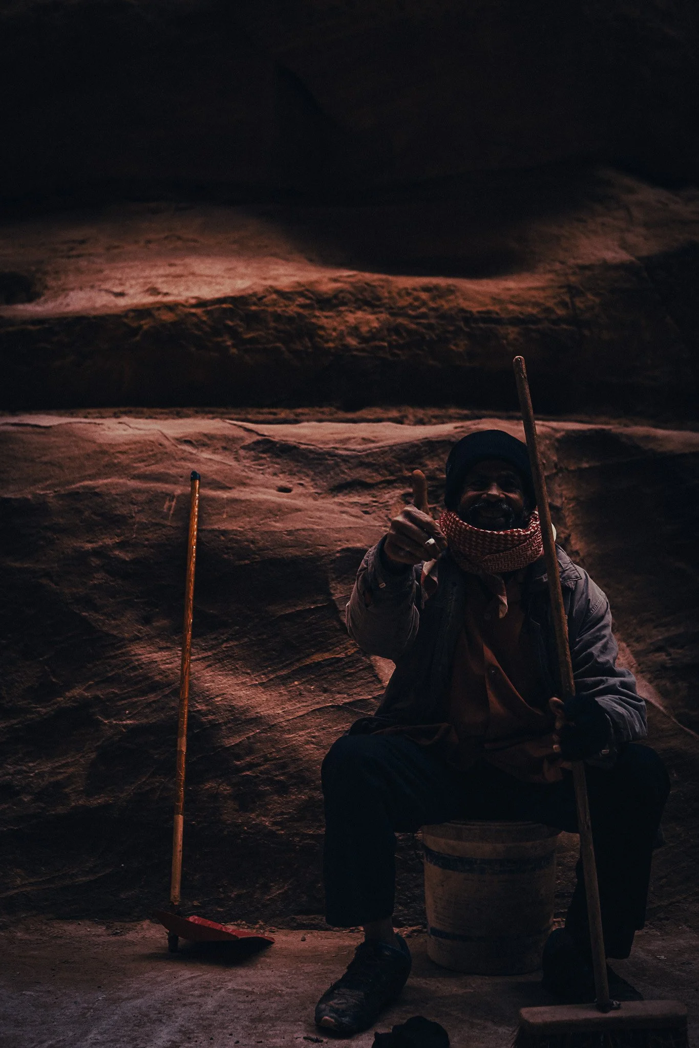 Bedouin man sitting against Petra’s sandstone rock, holding a walking stick and pointing toward the camera.