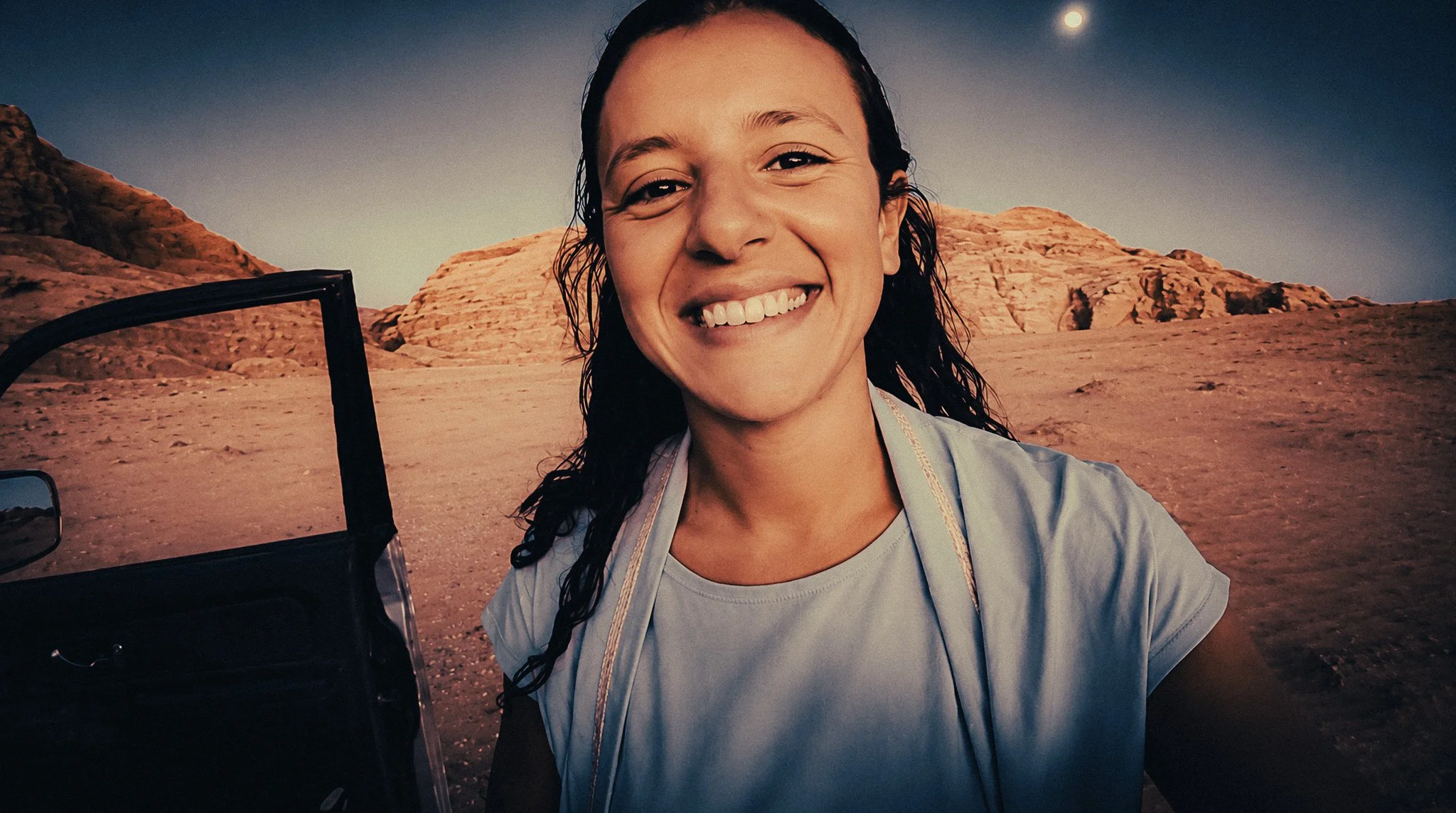 Woman smiling beside an open jeep in the Wadi Rum desert at sunset, with sandstone mountains and the moon behind her.