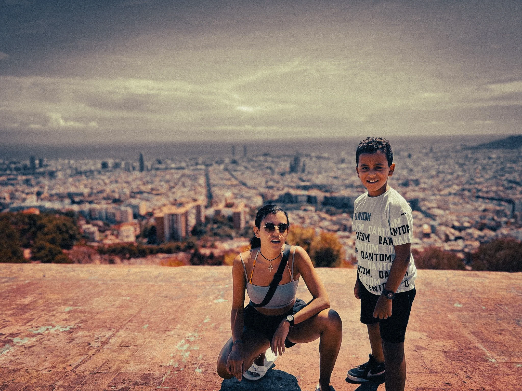 Woman and child overlooking Barcelona from a hill viewpoint, with the city skyline and Mediterranean coast in the background.