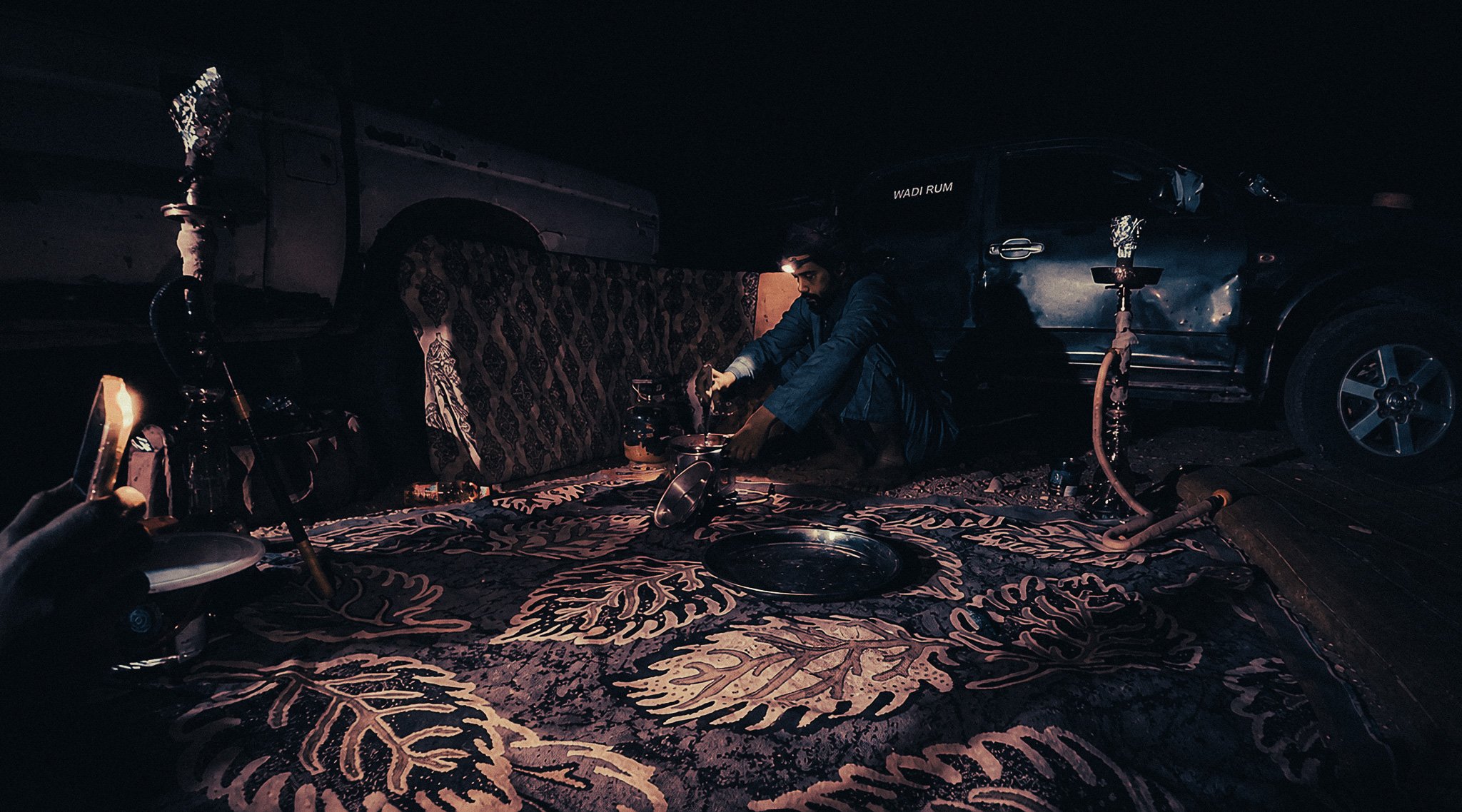 Bedouin man preparing food at a nighttime desert camp in Wadi Rum, surrounded by rugs, jeeps and shisha pipes.