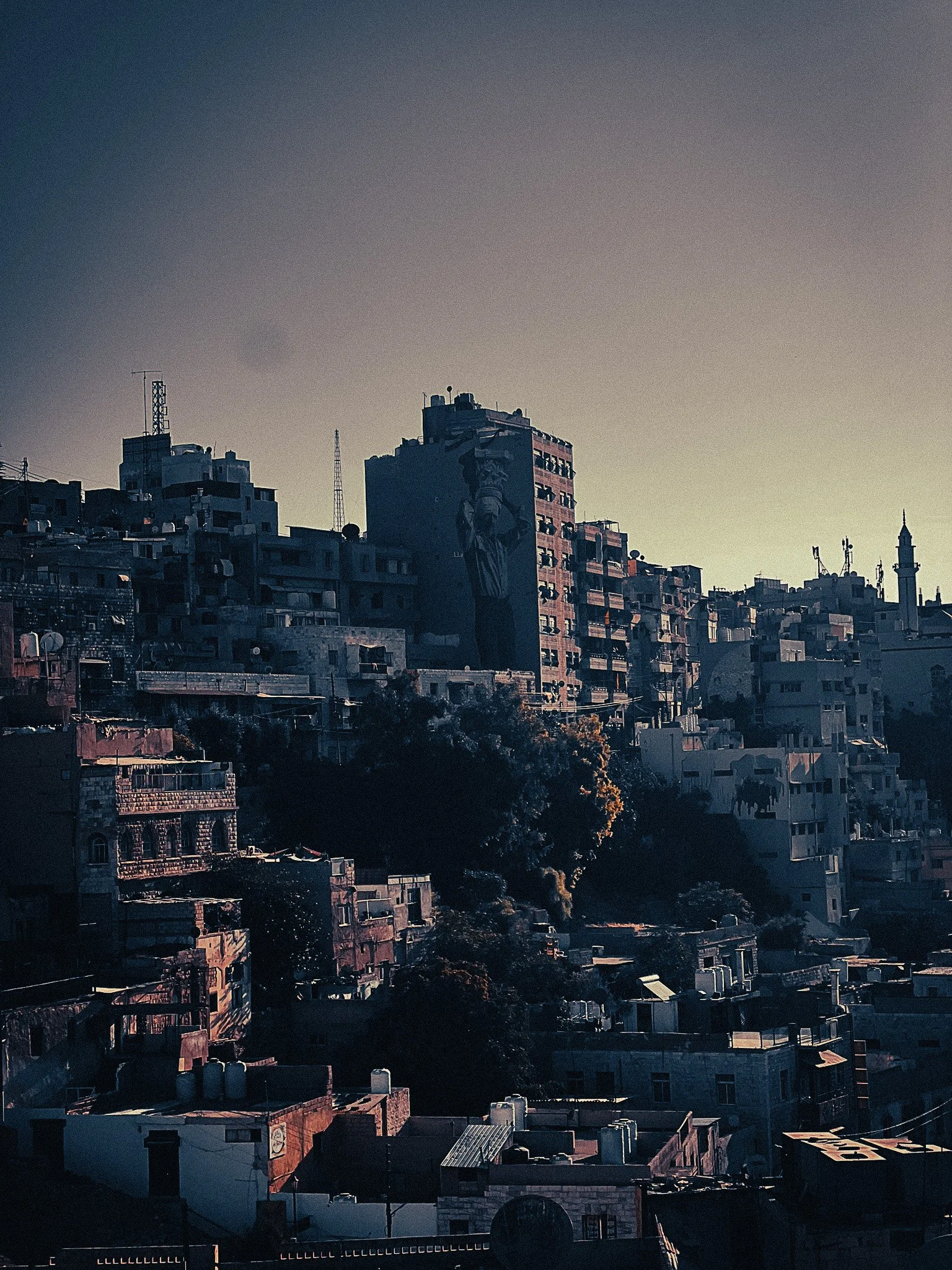 View over downtown Amman at golden hour, with stacked hillside buildings, shadowed rooftops, and the dense urban streets near the Roman Theatre area.