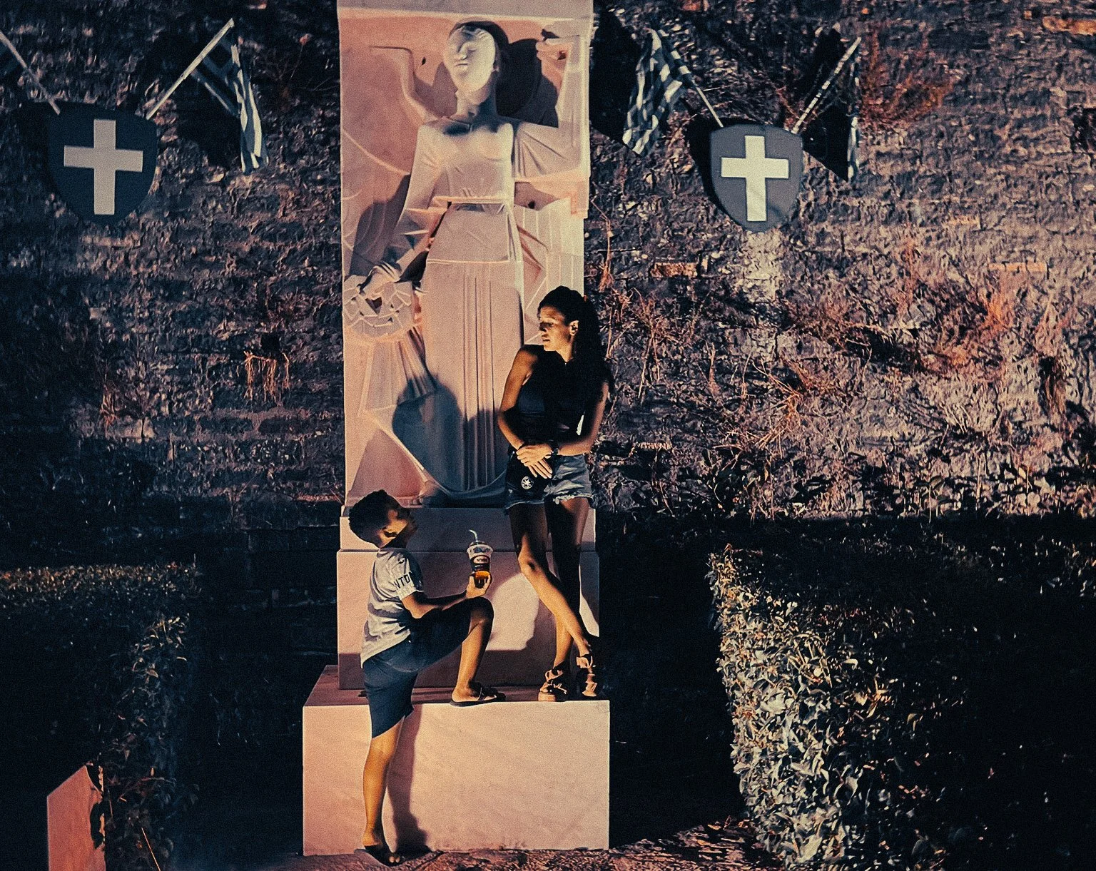 Mother and son standing beside a lit statue at night in Preveza Greece during a slow Mediterranean summer trip.