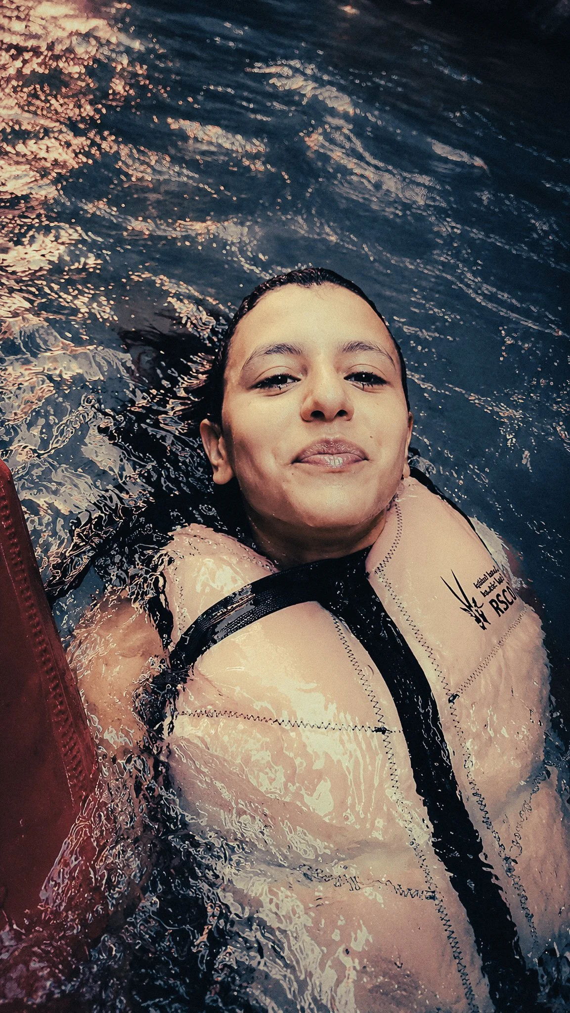 Woman floating on her back in Wadi Mujib wearing a life jacket, surrounded by dark blue-green canyon water.