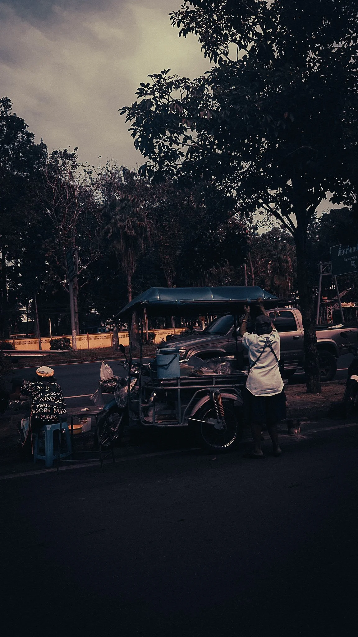 Street food cart and motorbikes in Khao Sok village, Thailand, with a vendor preparing his stall under roadside trees near the national park area.