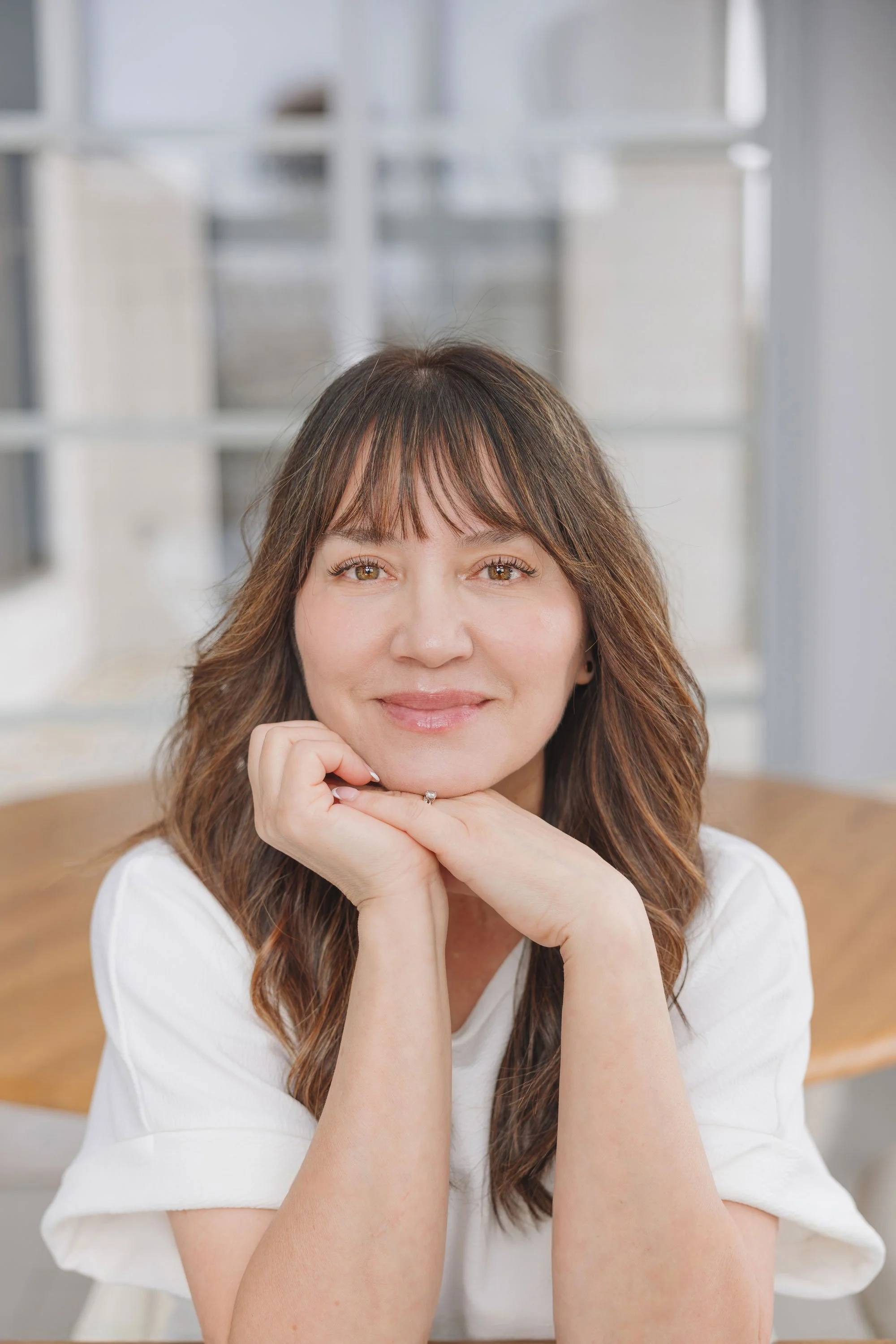 Portrait d'une femme souriante avec les cheveux bruns, assise à une table dans un intérieur lumineux.