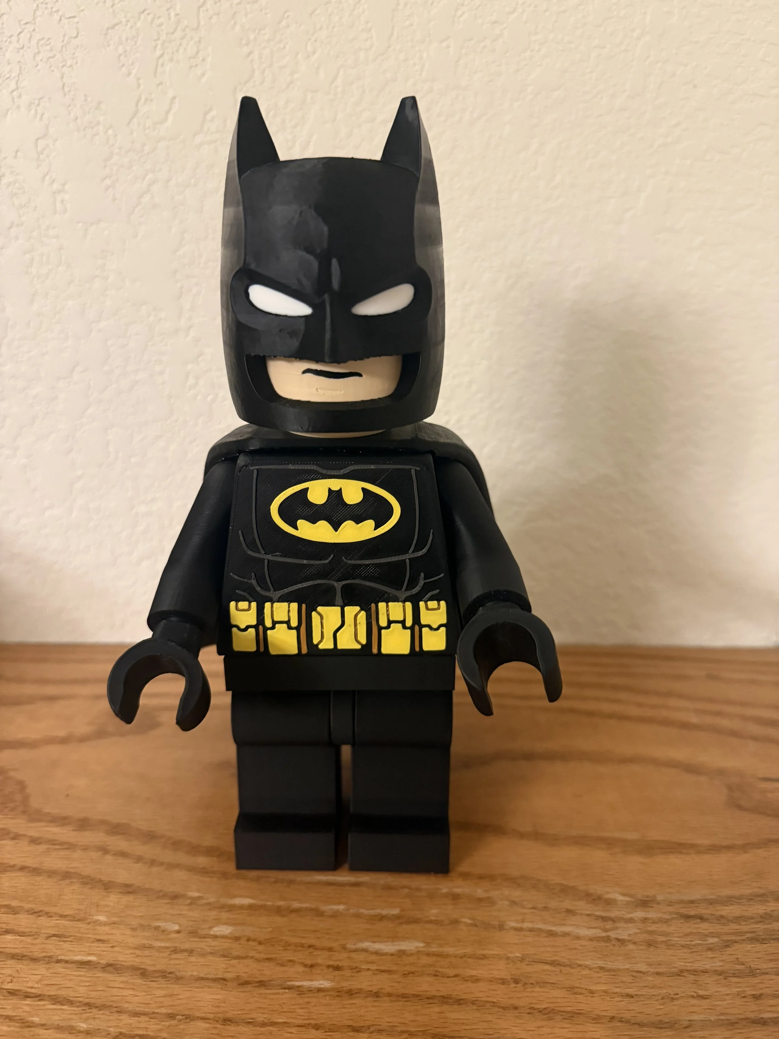 Lego Batman figurine with a black mask and costume, yellow Batman logo on chest, and yellow belt, standing on a wooden surface against a beige wall.