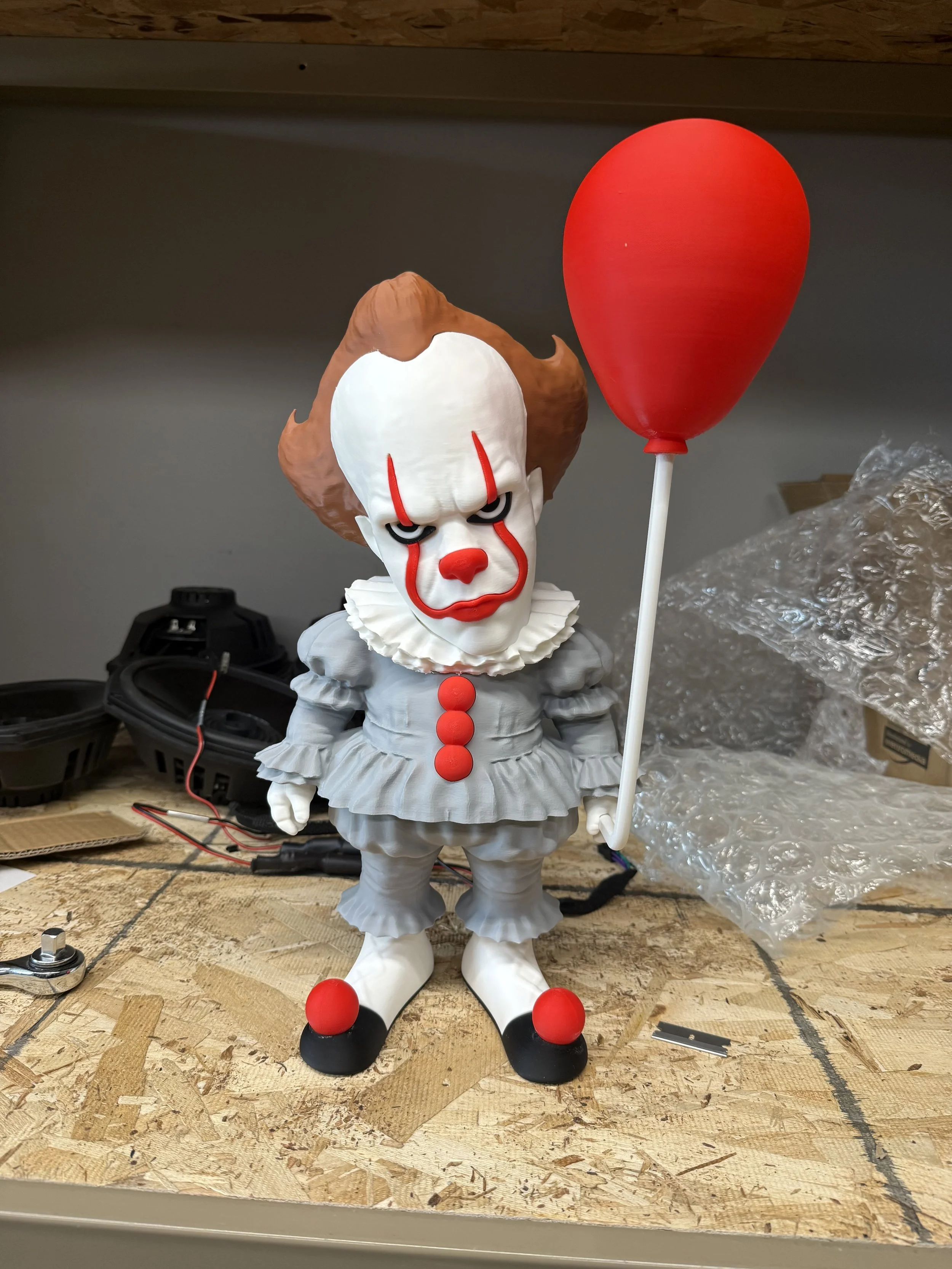 A figure of Pennywise the clown holding a red balloon, with a white face, red nose, red makeup, and a clown costume, stands on a workbench with tools and packaging around.