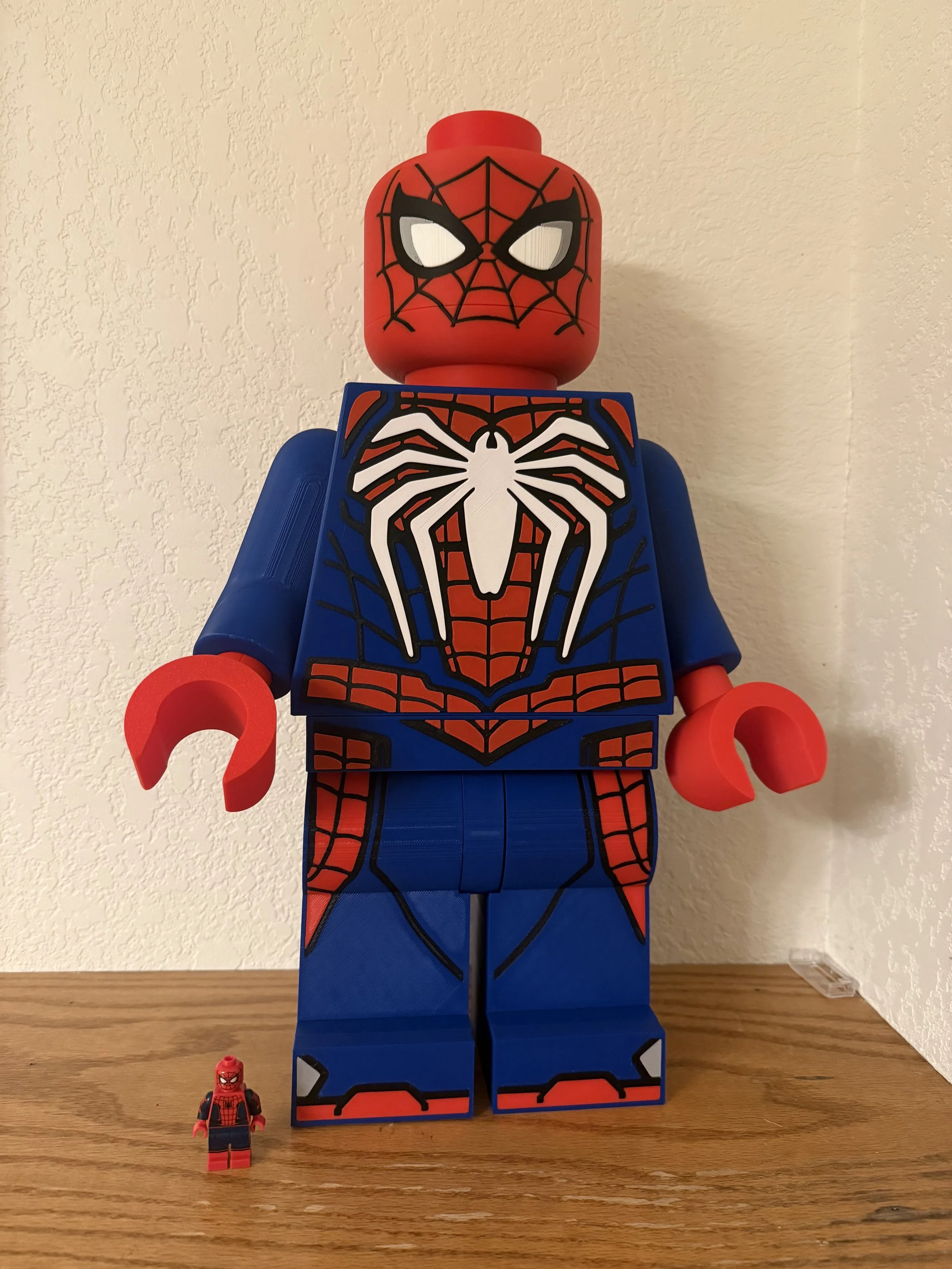 Large LEGO figure of Spider-Man standing on a wooden surface against a beige textured wall, with a smaller LEGO Spider-Man figure in front.