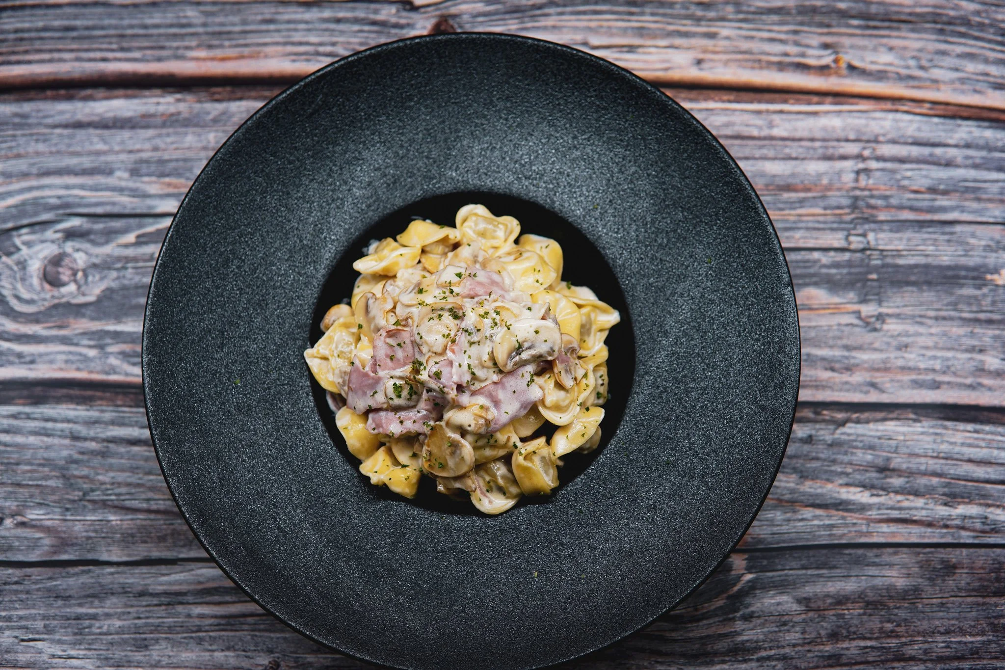 A black plate of creamy pasta with ham, mushrooms, and herbs on a rustic wooden table.