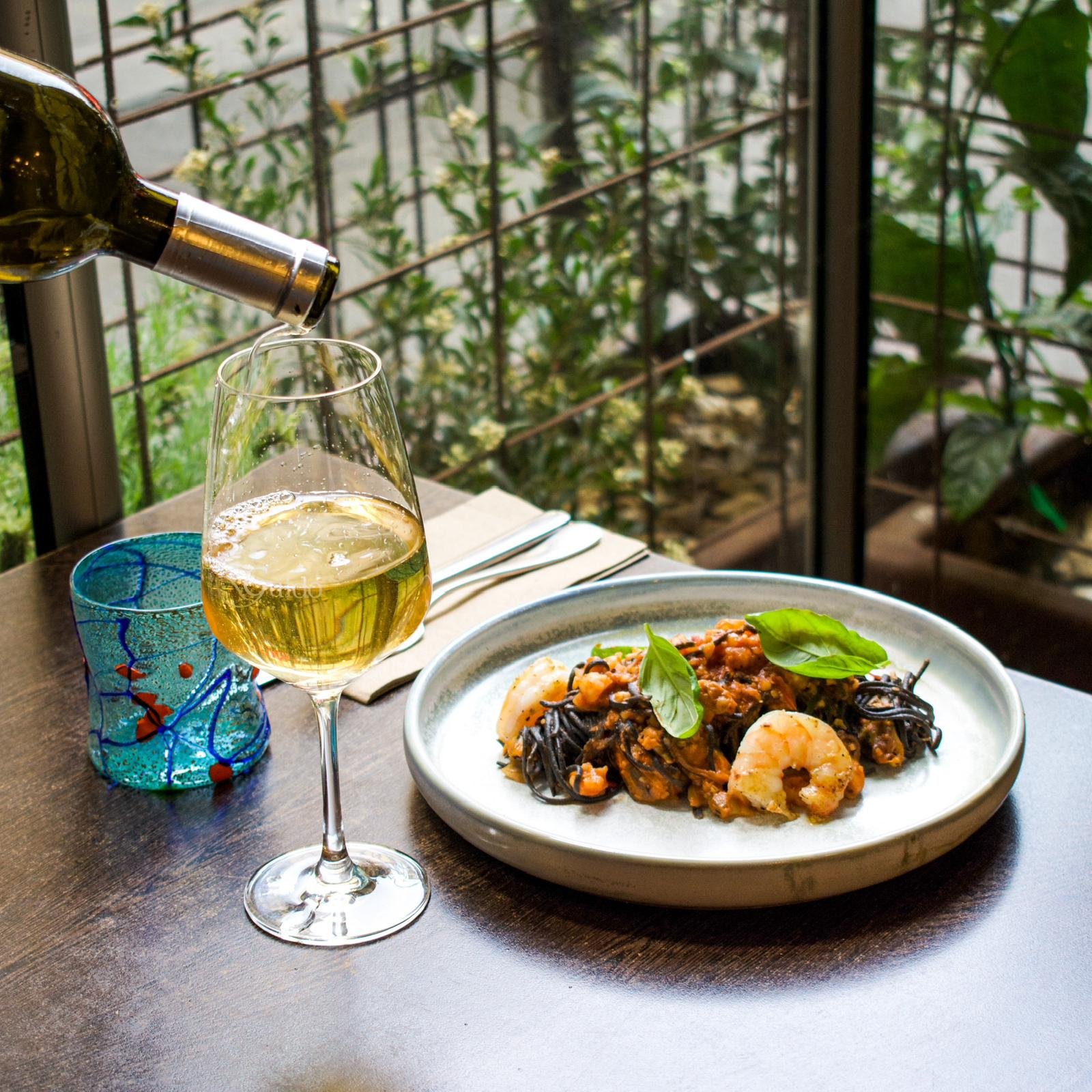 A glass of white wine, a blue decorative glass, and a plate of seafood pasta garnished with basil are on a wooden table at a restaurant with a green foliage background.