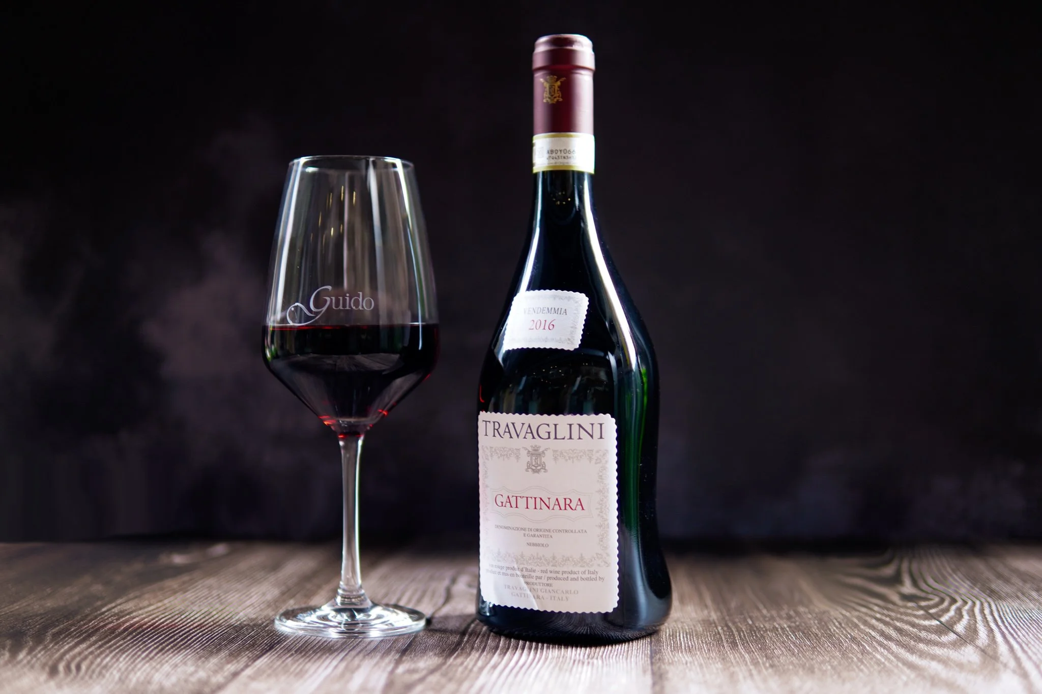 A bottle of Travaglini Gattinara red wine next to a glass filled with red wine on a wooden surface with a dark background.