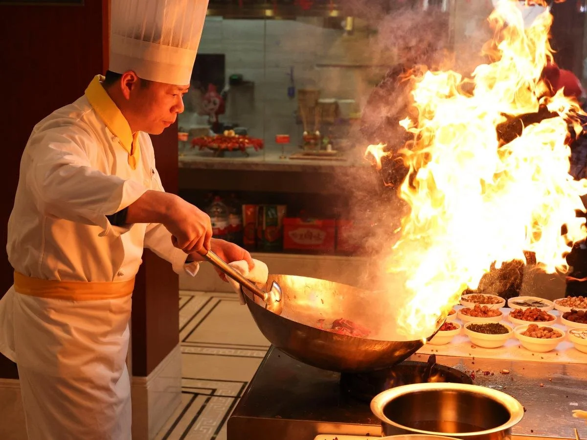 Koch in einer professionellen Küche, der beim Braten eines Gerichts in einer großen Pfanne eine Flamme verursacht.