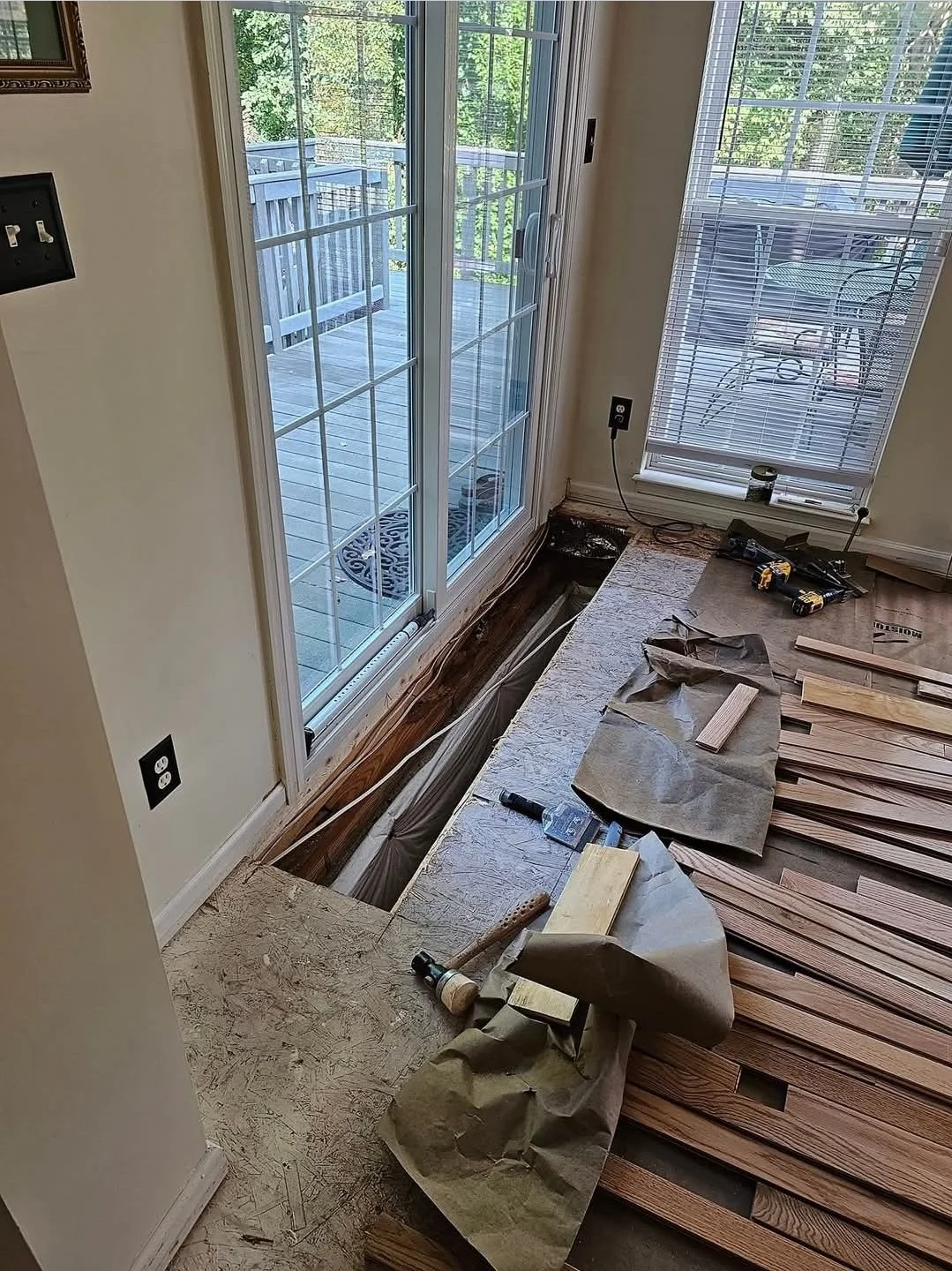Room under renovation with a sliding glass door leading to a deck, partially removed flooring revealing subflooring, and woodworking tools and materials scattered around.